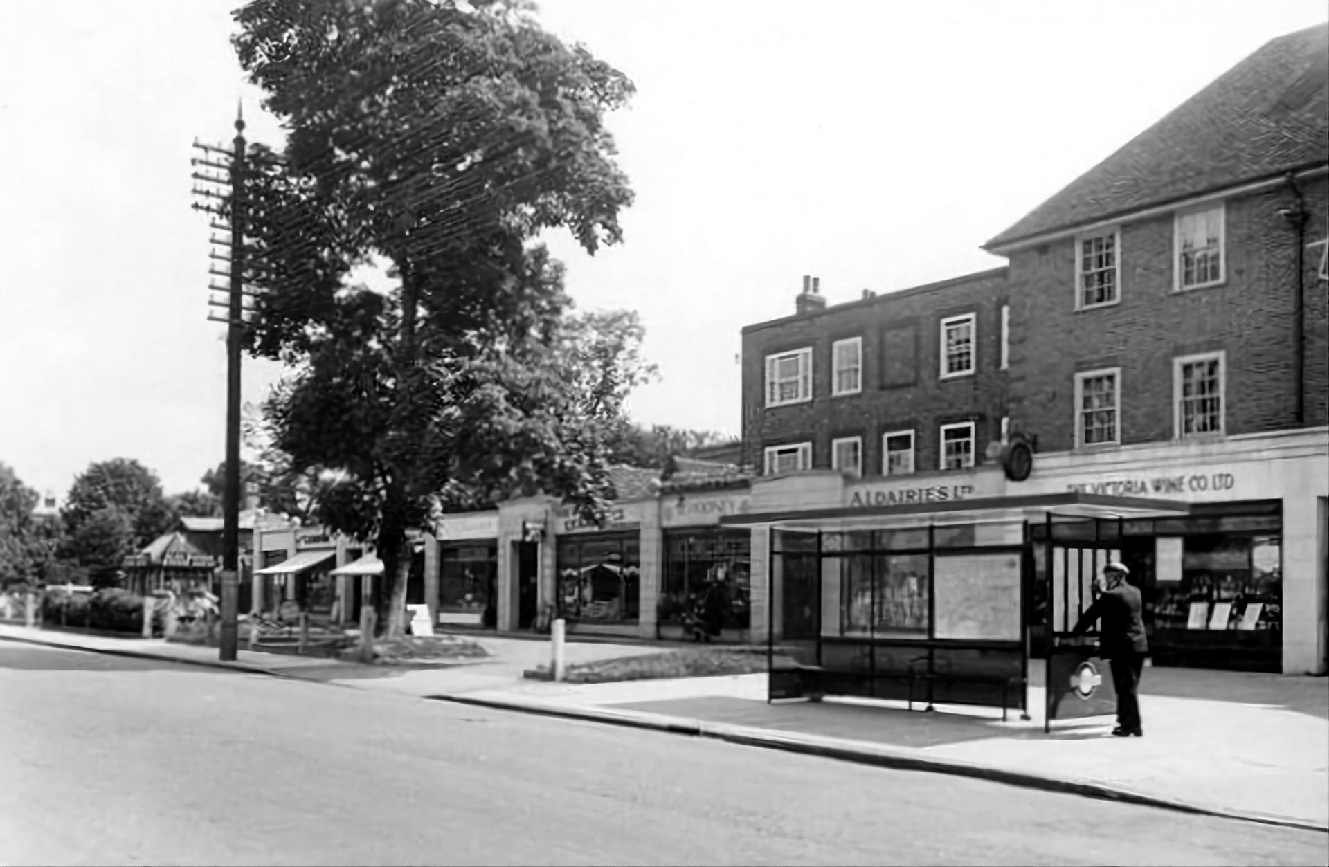 1933 "The Shops opposite the Bus Garage. Victoria Wine &amp; A1 Dairies Shop". Lloyd Rich identifies this as 1933 "showing the new logo for the newly formed London Passenger Transport Board who were formed on July 1st 1933 - this is a posed photo showing one of their new bus shelters and shows their original logo where the man is standing." David Crane comments "I remember the dairy round behind the shops very well. My father was a milkman for them in the early sixties. Amazing electric milk floats so far advanced in those days." Keith Whitfield adds "My father was also a milkman there, he did Bentley Heath, Waggon Road and Crews Hill". Veronica Turner says "My uncle Alf Hill delivered the milk to St John's school in 1954-6 when I was there, probably before." (Courtesy Jill Hellary)