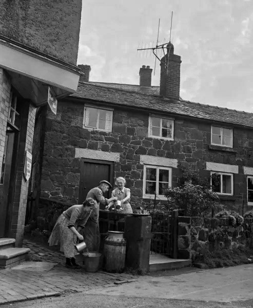 1956 Ruyton-XI-Towns in time of water shortage. Churns are placed outside each house and are filled with ten gallons of water twice a week from a water cart. To augment their ration of churn water villagers place baths and other receptacles under drainpipes to catch water for washing and bathing. John Beegles, 74, getting water from the churn outside his house for his 74-year-old wife Alice. His next door neighbour, Mrs Betty Guyatt, 31, also takes her share. (Courtesy Mirrorpix)