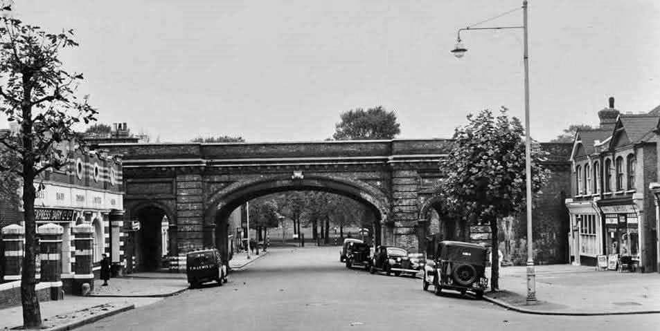 1954 Herne Hill Depot and shop, with TH Lewis van. William D.T. Davis comments "Looking North down Rosendale Road towards the beautifully built 1869 railway bridge that carries the lines between Tulse Hill and Herne Hill Stations. To the immediate left of frame are Edwardian built Peabody Estate Buildings; naked of the iron railings pointlessly removed as a propaganda exercise at the start of the war and like the bulk collected through out the country, quietly dumped following peace into the Thames Estuary. Alongside the Estate was the Express Dairy, whose obviously notable smell passing by was the daily combination of the hosing down of spilt milk with water. The Dairy shop in the front, although busily used by shoppers, was closed in about 1970. To the right is the corner with Gurnsey Grove. Under the railway bridge, on the left hand corner with Norwood Road, was the corner tobacconist/newsagents, post office and pillar box. When it closed in the mid 1970s, the post office moved business to the middle of the parade of shops opposite Peabody Estate and was run by Mr &amp; Mrs Flanagan; who had formally run their sweet/newsagents shop a few doors up, one unit away from the corner with Hawarden Grove." (Courtesy William D.T. Davis)