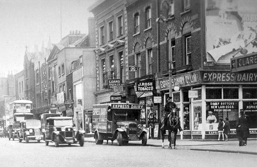 1936 A detail from a wonderful, commercial postcard view of Hammersmith Road, from around 1936. The shop assistant in the Express on the corner can be seen in her ‘whites and mop cap’, while two school girls peered into the window. They were probably pupils from the Sacred Heart High School across the road, which still survives. New laid eggs and cream are advertised while the delivery driver (spot his legs through the chassis!) sorted his load. This Ford ‘AA’ 30cwt box van, Express fleet no 25, GW 9102, was supplied on 8/12/31 by AE Gould Ltd, for £165 8 6d. TH Lewis built the body, which came complete with roller shutters front and rear and cake trays, for £155 19s. Chassis no AA4793085 was built at the new Dagenham plant during the first month of production there. The first vehicle off the line on 1/10/31 was an ‘AA’ truck, on chassis no AA4791110. The van was sold to G Ridler of London W11, in December, 1942, for £27 10s. Apart from the mounted policeman, passing traffic includes a large Sunbeam, an Austin 16/18hp and Morris Oxford. (Courtesy The Express Dairy Motorised Fleet, Allan Bedford, Heritage Machines)