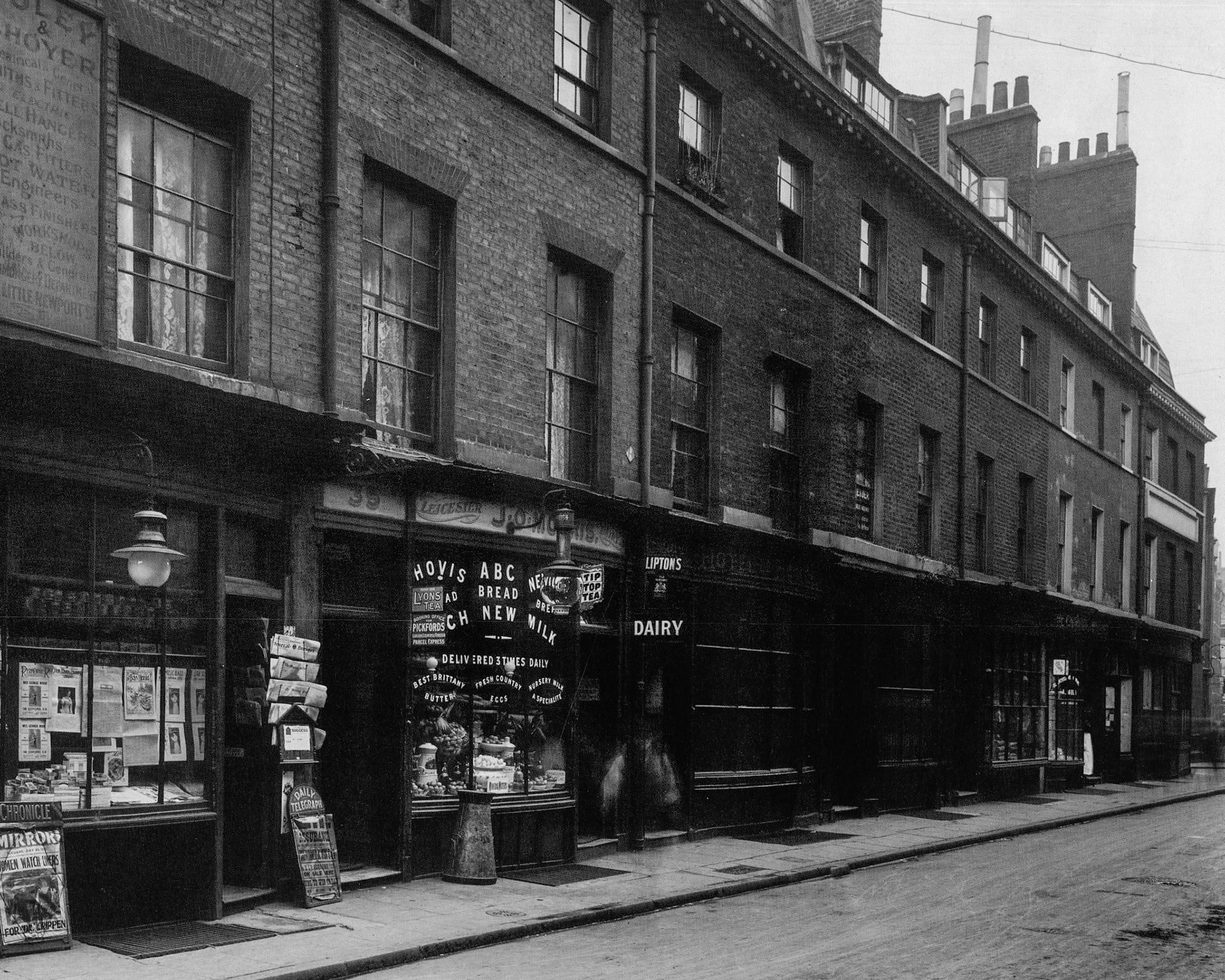 1910. 34-39 Lisle Street, Soho, terrace of late 18th century houses with contemporary shopfronts and a wealth of detail. J.O. Morris, Leicester Dairy advertises 'New Milk' for sale. The newsagents on the left has an enamel sign for the National Telephone Company to advertise a public telephone, and the placard for the 'Mirror' leads with 'Women Watch Liners for Dr Crippen'. (Courtesy GLC Historic Buildings Collection)