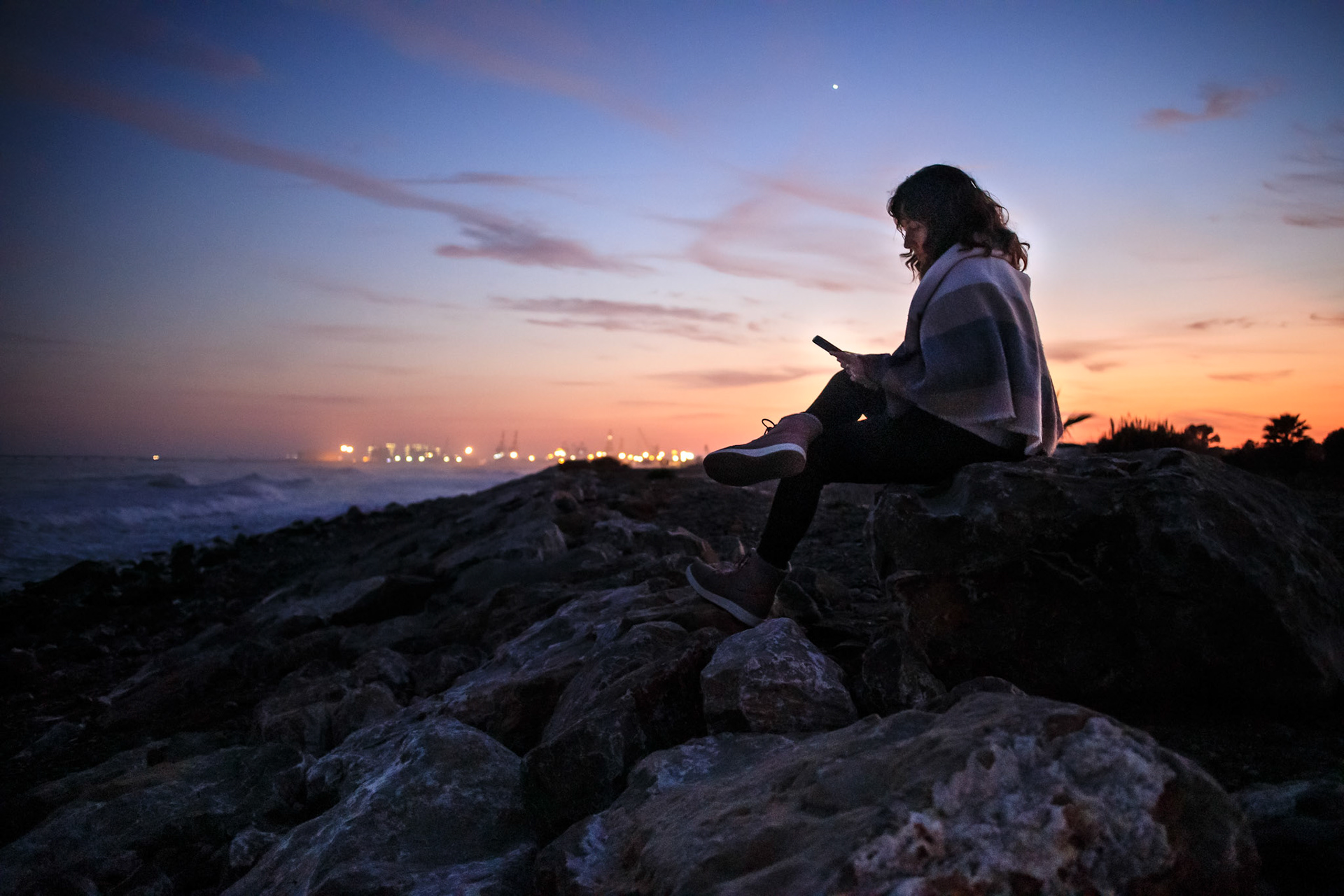 Adult woman on a pebble beach in winter enjoying with the cell phone at the shore of the beach at sunset