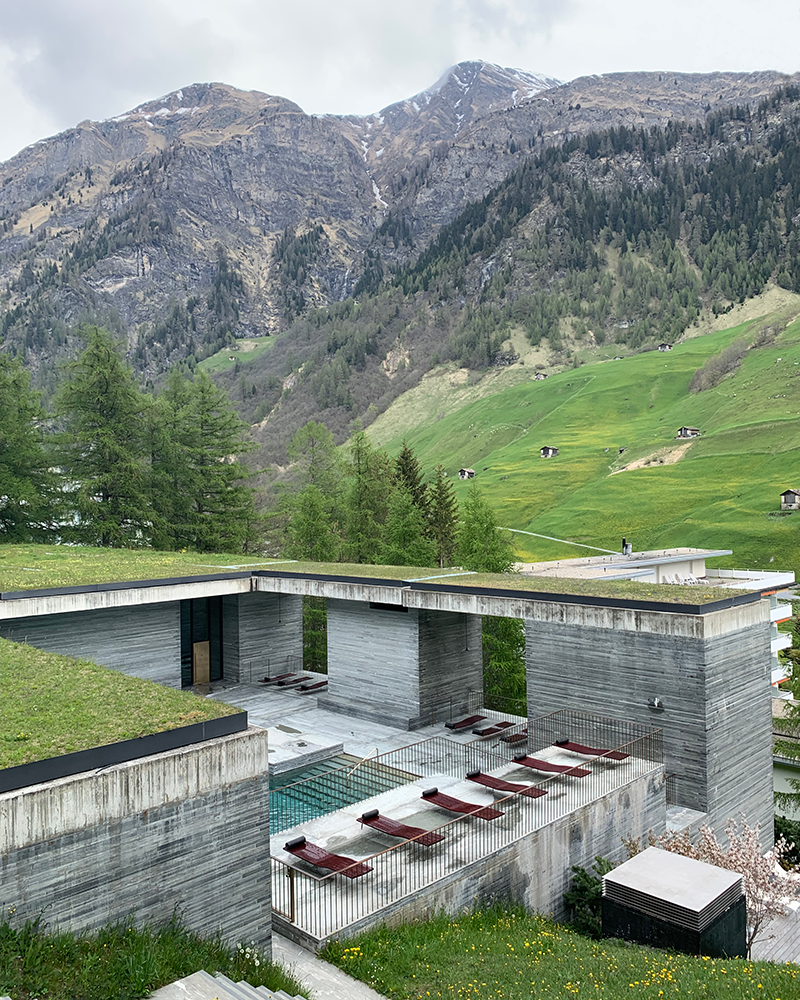 Therme Vals Exterior Pool and lounge chairs by Peter Zumthor with green roof in front of tall pine trees and a snow-capped mountain with traditional swiss farm huts on the other side of the valley