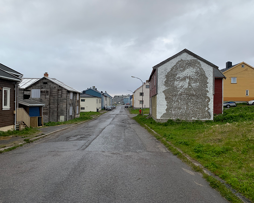 Mural by Vhils in Vardø, Norway the the side of a house on an empty wet residential street under a cloudy sky