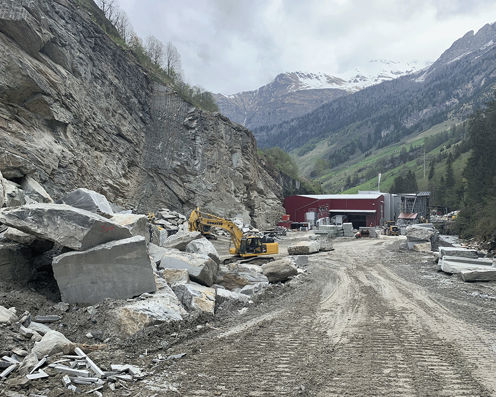 Truffer AG Quarry in Vals, Switzerland with a yellow excavator and red saw mill in front of snow-capped mountains and blocks of Valser stone