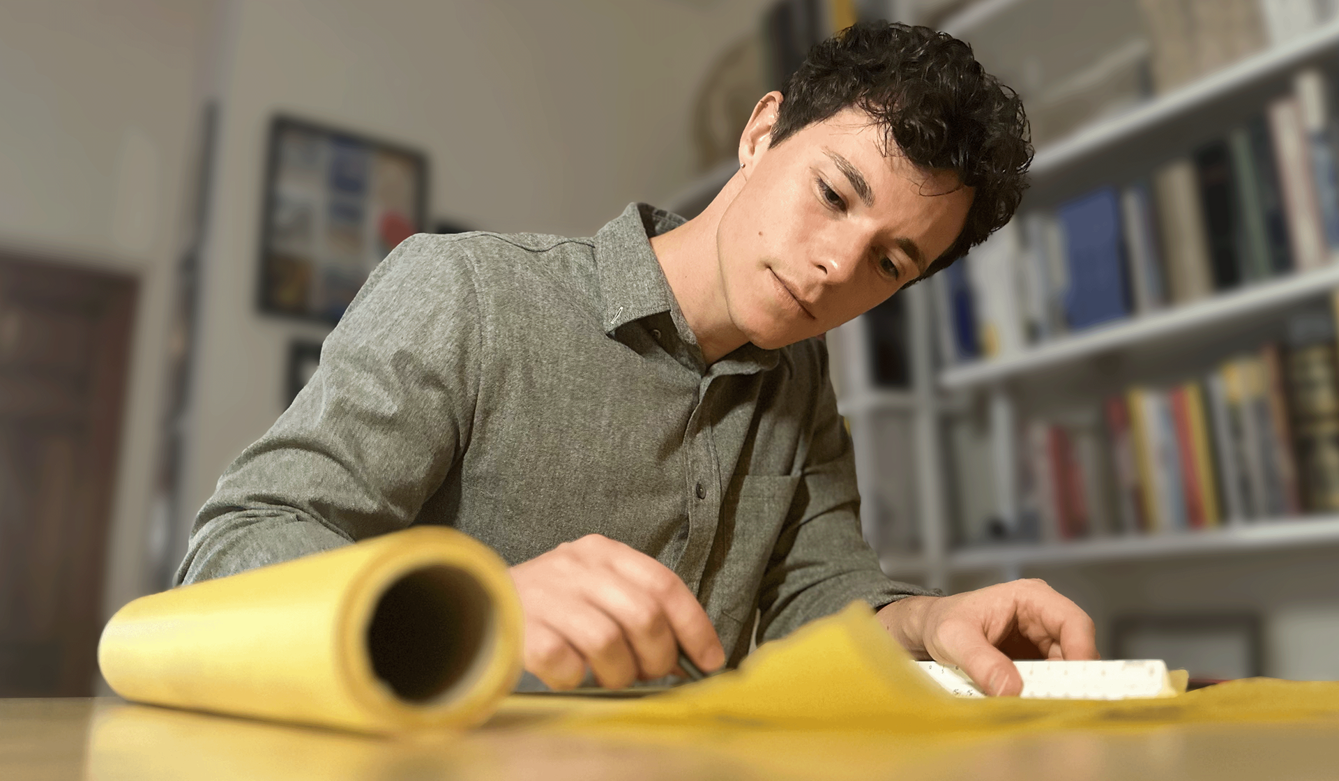 David Sweere, AIA, NCARB sketching with a ln architectural scale and charcoal pencil on a roll of yellow trace paper on a wood desk in front of a book shelf and architectural models.