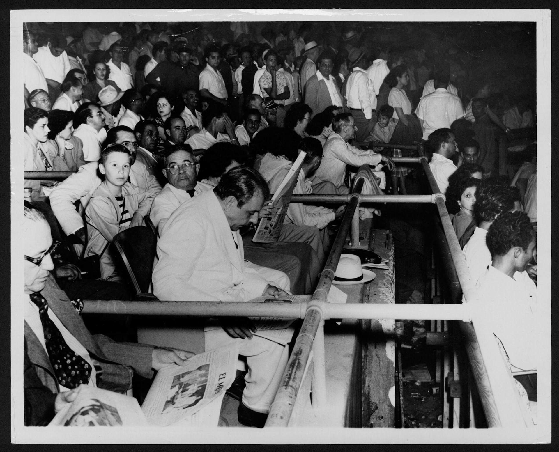 LUIS MUÑOZ MARÍN SENTADO ENTRE LOS ESPECTADORES DEL JUEGO DE PELOTA ENTRE SANTURCE Y PONCE MIENTRAS LEE UN PERIÓDICO.PARQUE SIXTO ESCOBAR.FEBRERO 11, 1949