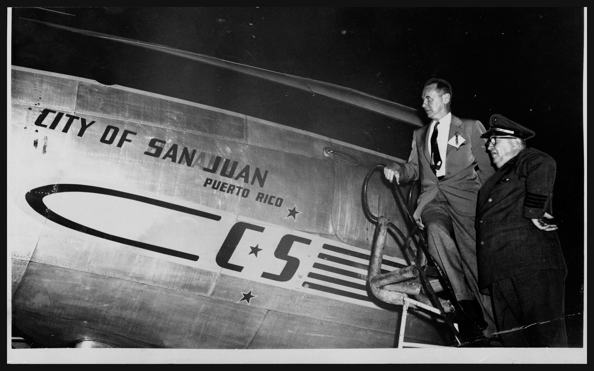 Pie de la foto: "El señor Sydney A. Stewart, presidente de la línea Chicago &amp; Southern, y el capitán del avión City of San Juan, Ben Catlin, a su arribo a San Juan el sábado por la noche al inaugurarse el nuevo servicio entre Nueva Orleans y San Juan. Catlin es uno de los diez más antiguos pilotos conocidos, pues tiene 37 años en el servicio activo comercial. El avión, un Constellation de cuatro motores, trajo una nutrida representación de Luisiana con motivo del acontecimiento. El City of San Juan tiene pintadas banderas de Haití, Estados Unidos y la República Dominicana y se espera pronto aparezca la del Estado Libre Asociado."; Información: El Mundo, 12/enero/1953.Enero 12, 1953