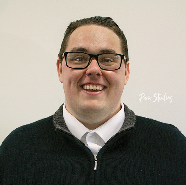 A portrait of Bertram, they wear a white shirt underneath a dark blue jumper and glasses.
