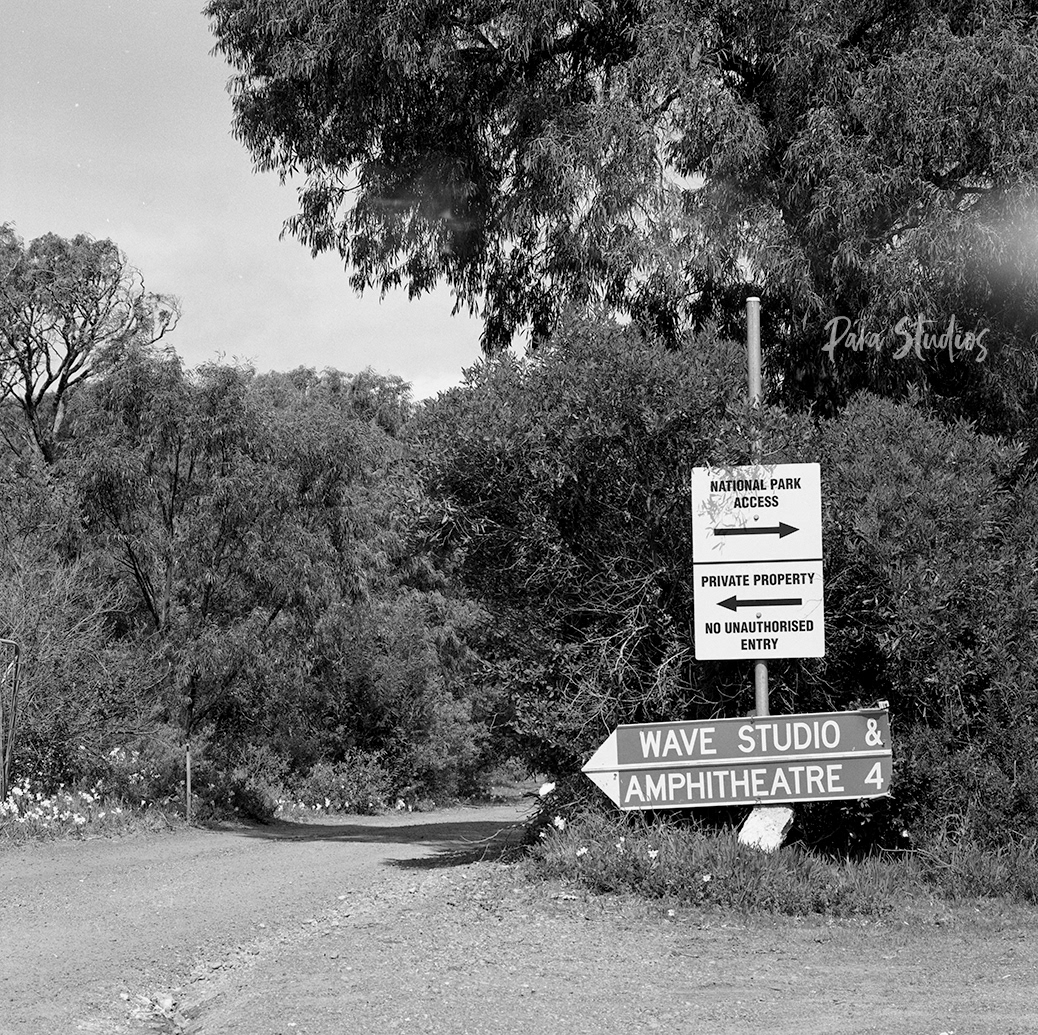 A black and white photo of a dirt driveway in the middle of the bush. To the right are signs which read, "National Park entrance", "Private property no unauthorised entrance", and "Wavehouse Studio & amphitheatre".