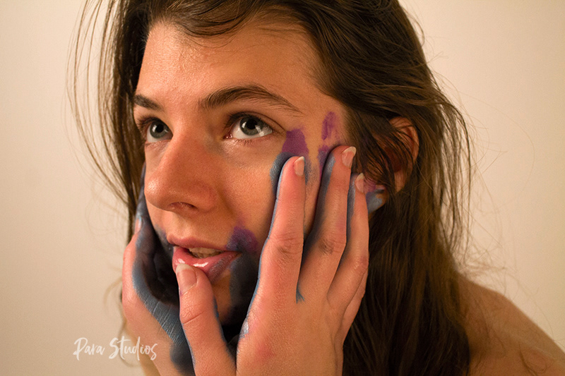 "Touched". A woman looks up as painted hands touch her face and pull down her lip. Paint is smeared across her face.