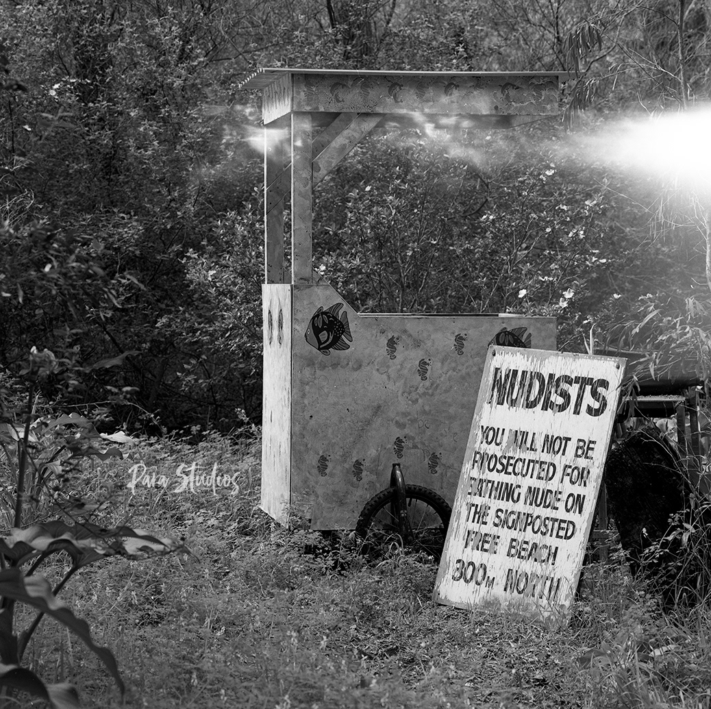 A black and white photo of a broken down cart and sign in the bush. The sign reads, "Nudists will not be prosecuted for bathing nude on the signposted free beach 300m north."