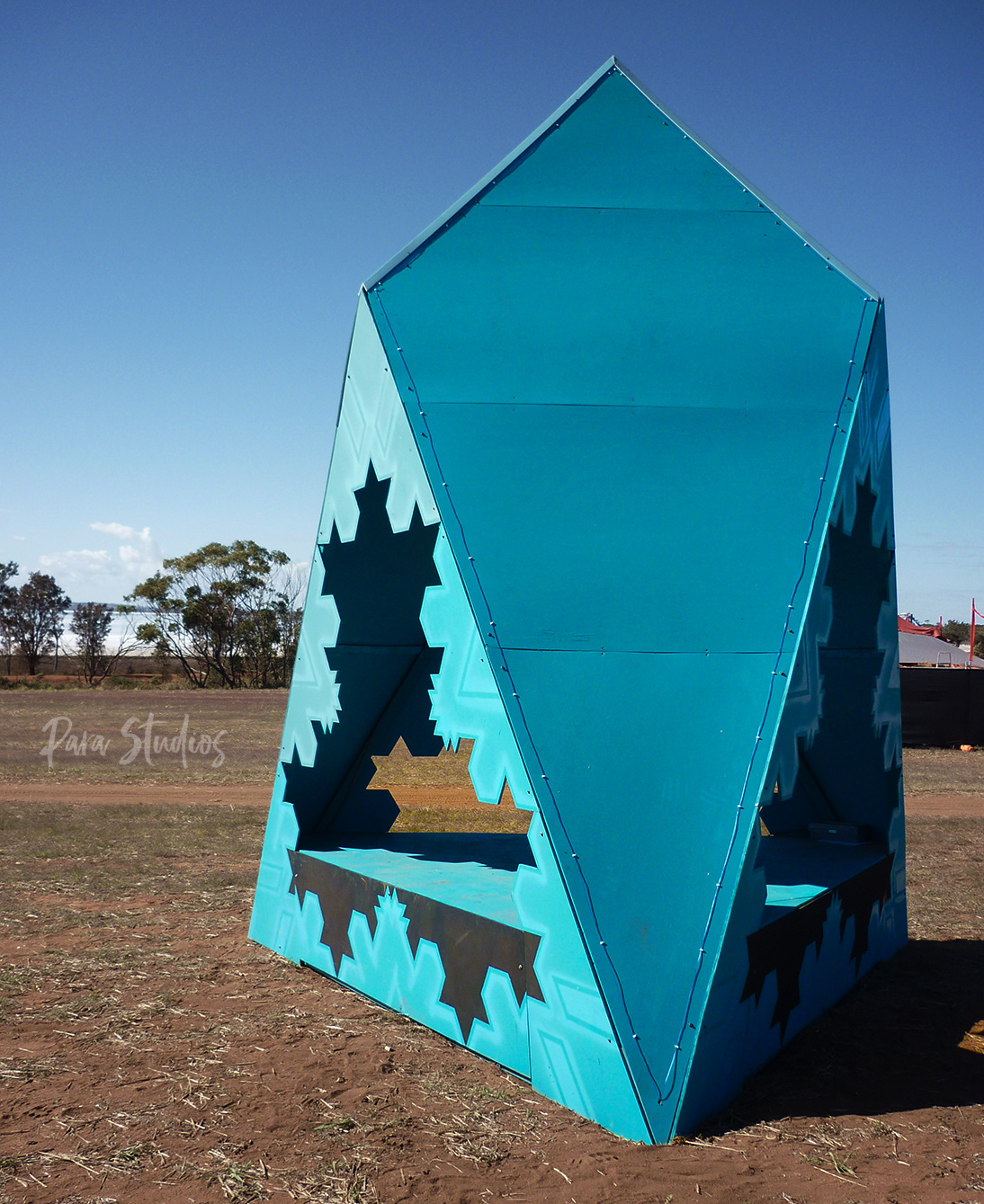In the middle of a paddock there stands a 2m tall art installation painted bright blue. The structure consists of 3 quadrilateral sides and 4 equilateral triangle sides, with a sitting platform in the middle. Behind the tree line you can see Jilakin salt lake.
