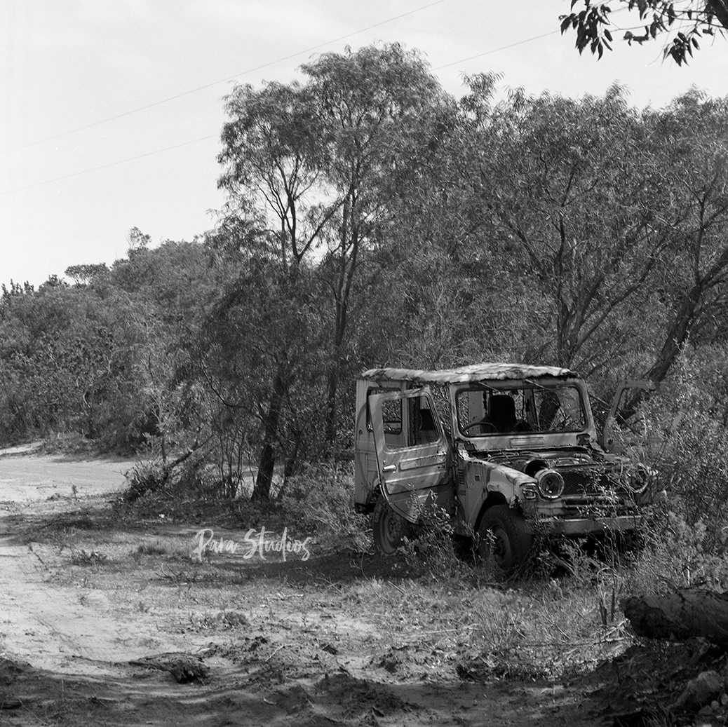 A black and white photo of a car crash in the middle of the bush. The car sits to the right of a sandy road, its body damaged, bonnet removed, and doors left wide open. Over head are trees and powerlines.