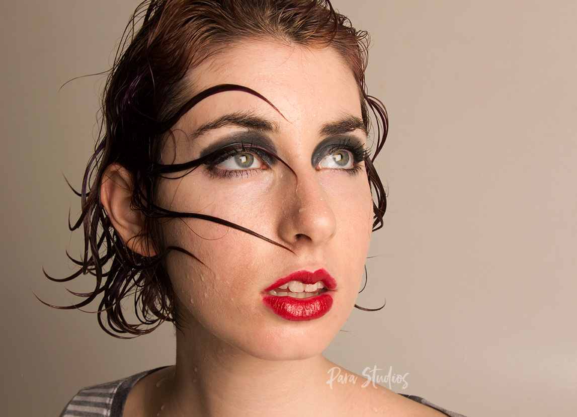 "Absorbed". A woman wears dark eye makeup and bright red lipstick. Her hair is wet and is sticking across her face as if she just got out of water.