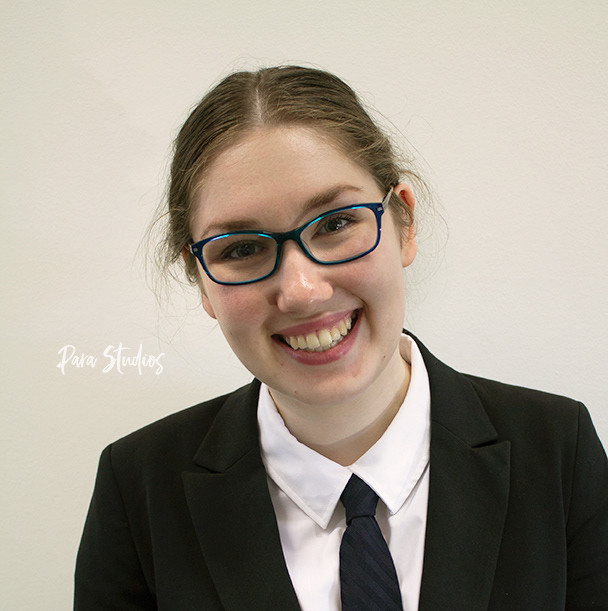 A portrait of Harriet, they wear a black jacket, black striped tie and glasses.