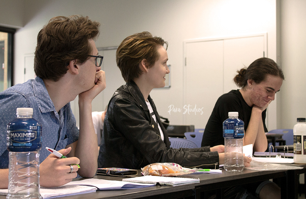 A shot of the show directors taking notes during a rehearsal. Wearing blue and black, they sit at a desk scattered with notebooks and snacks.