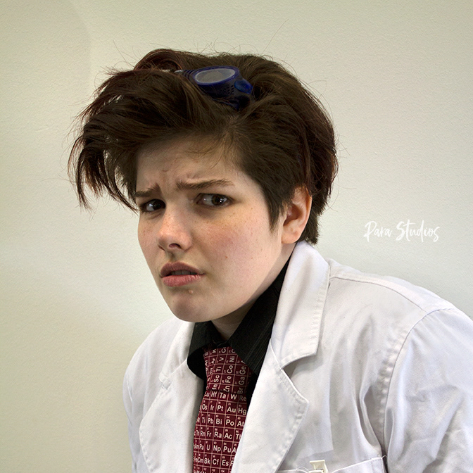A portrait of Alessandra, they wear a white lab coat, a red periodic table tie and goggles on top of their head.