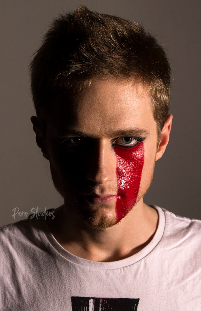 "Fierce". A man stares down the camera. Half of his face is lit with the other half in extreme shadows. He wears dark eye makeup and thick red lipstick which runs from the bottom of his eye down to his chin.
