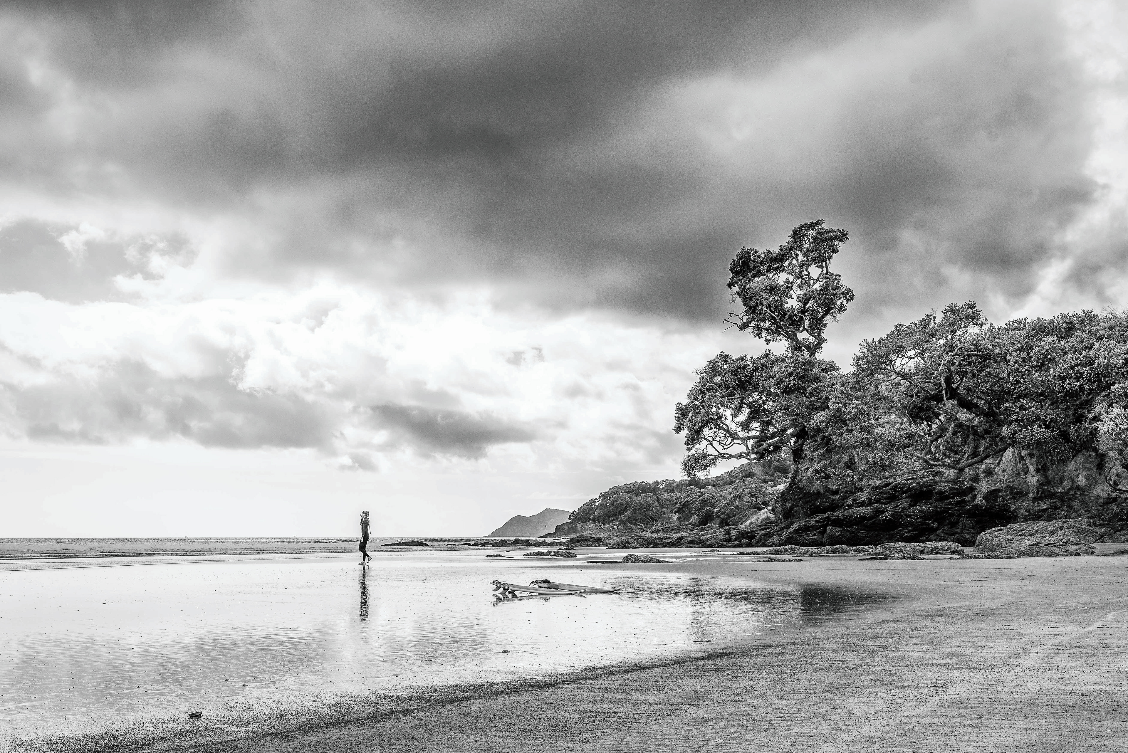 wetsuit clad surfer standing in the shallows with surfboards in the foreground, dramatic clouds in the background, in monochrome