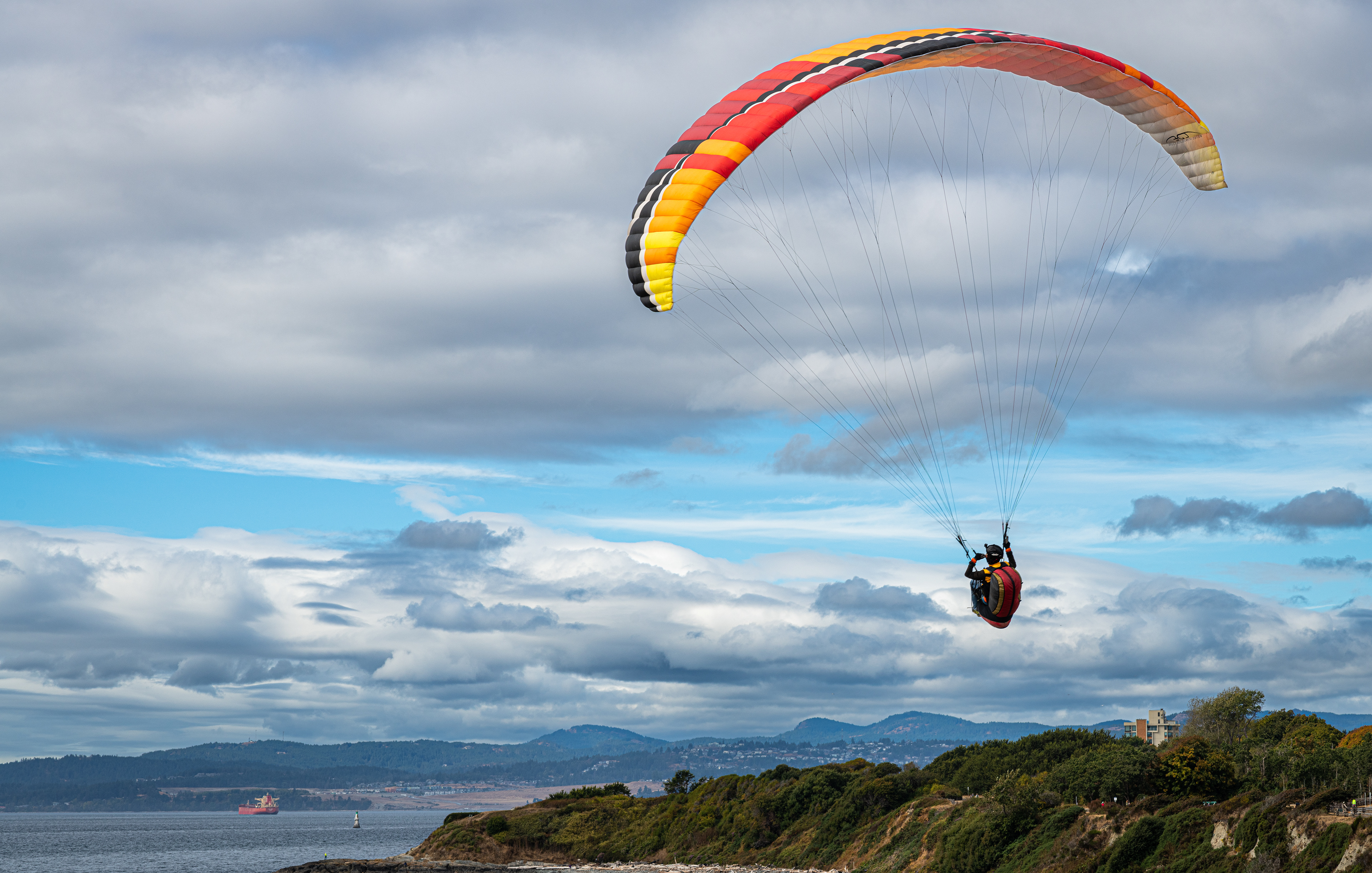 Brightly coloured parasailer against white cumulus clouds, above the ocean alongside of Beacon Hill Park, Victoria, BC