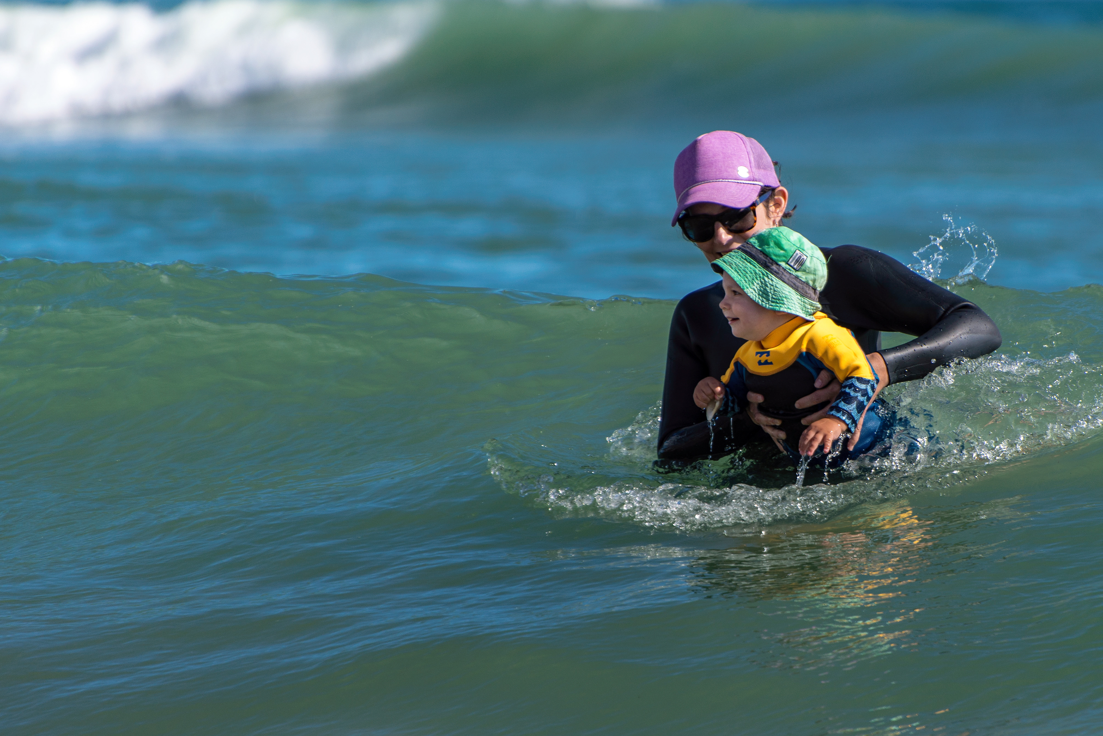mom with purple ball cap standing chest deep in the waves holding her infant son dressed in wetsuit and sunhat at Papamoa, NZ