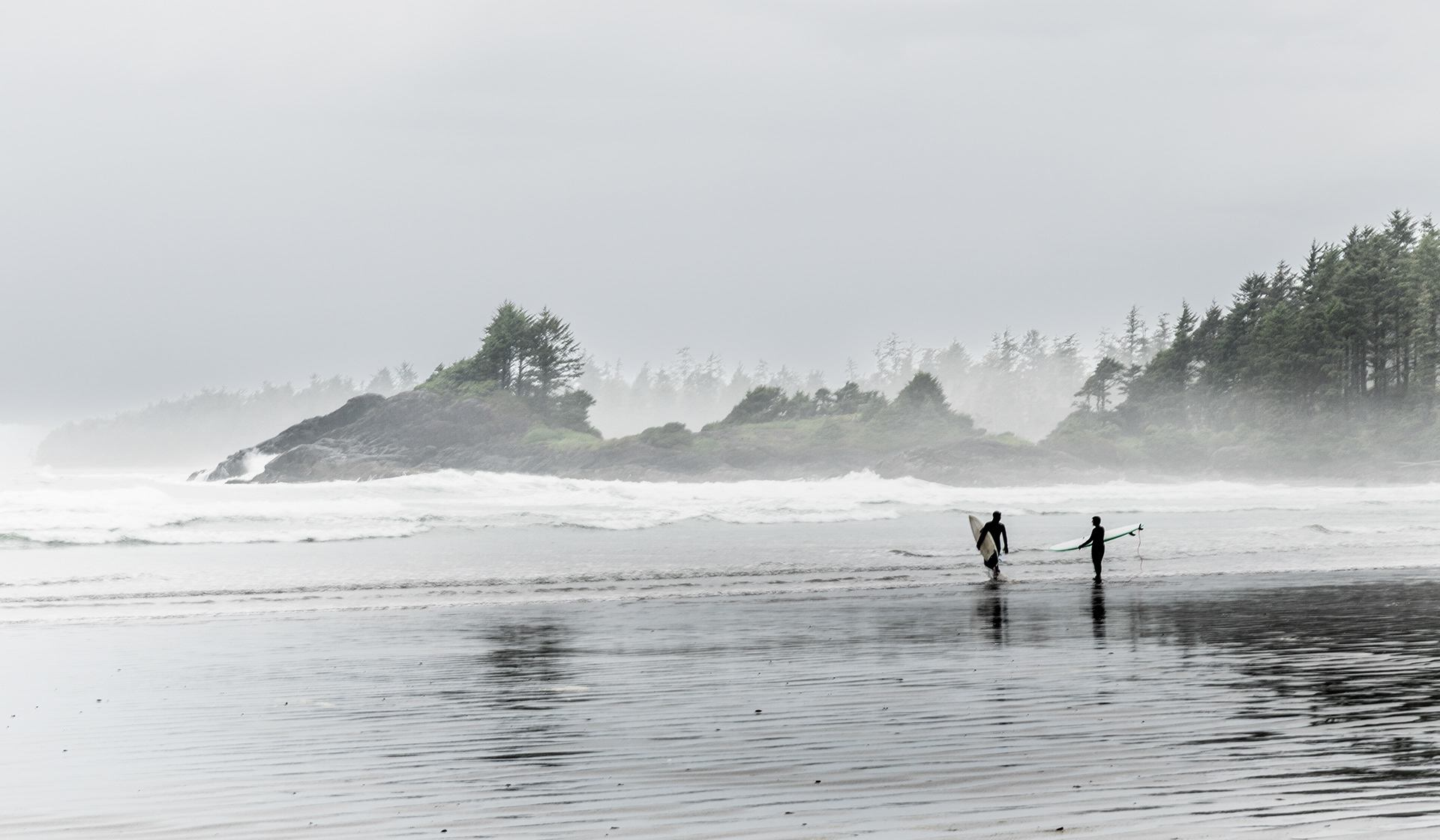 two wetsuit clad surfers with boards wading into the winter surf at Cox Bay, Tofino, BC, with fog shrouded rocky headlands in the background