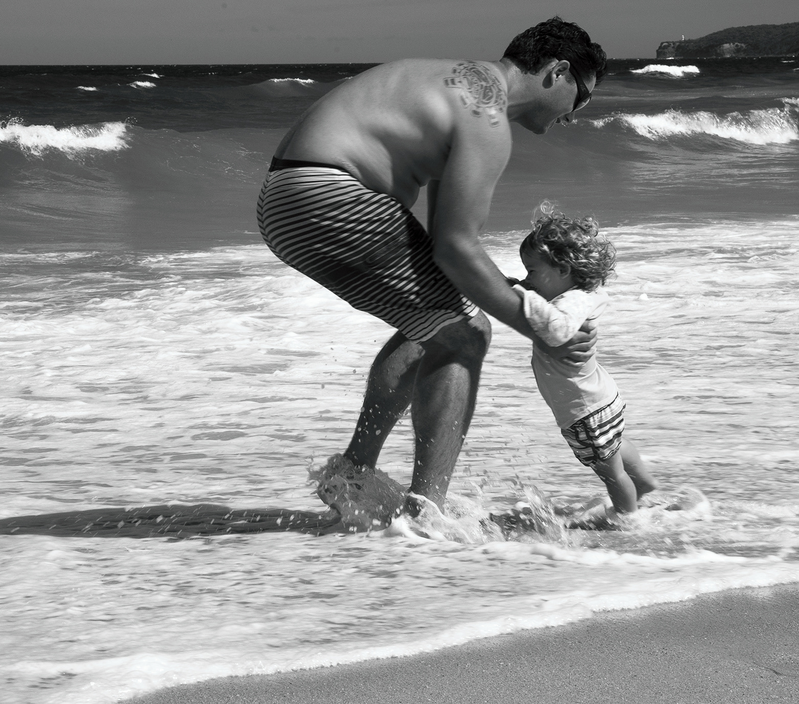 Faher picking up a infant at surfs edge with white capped surf in the background, near Ulladulla NSW