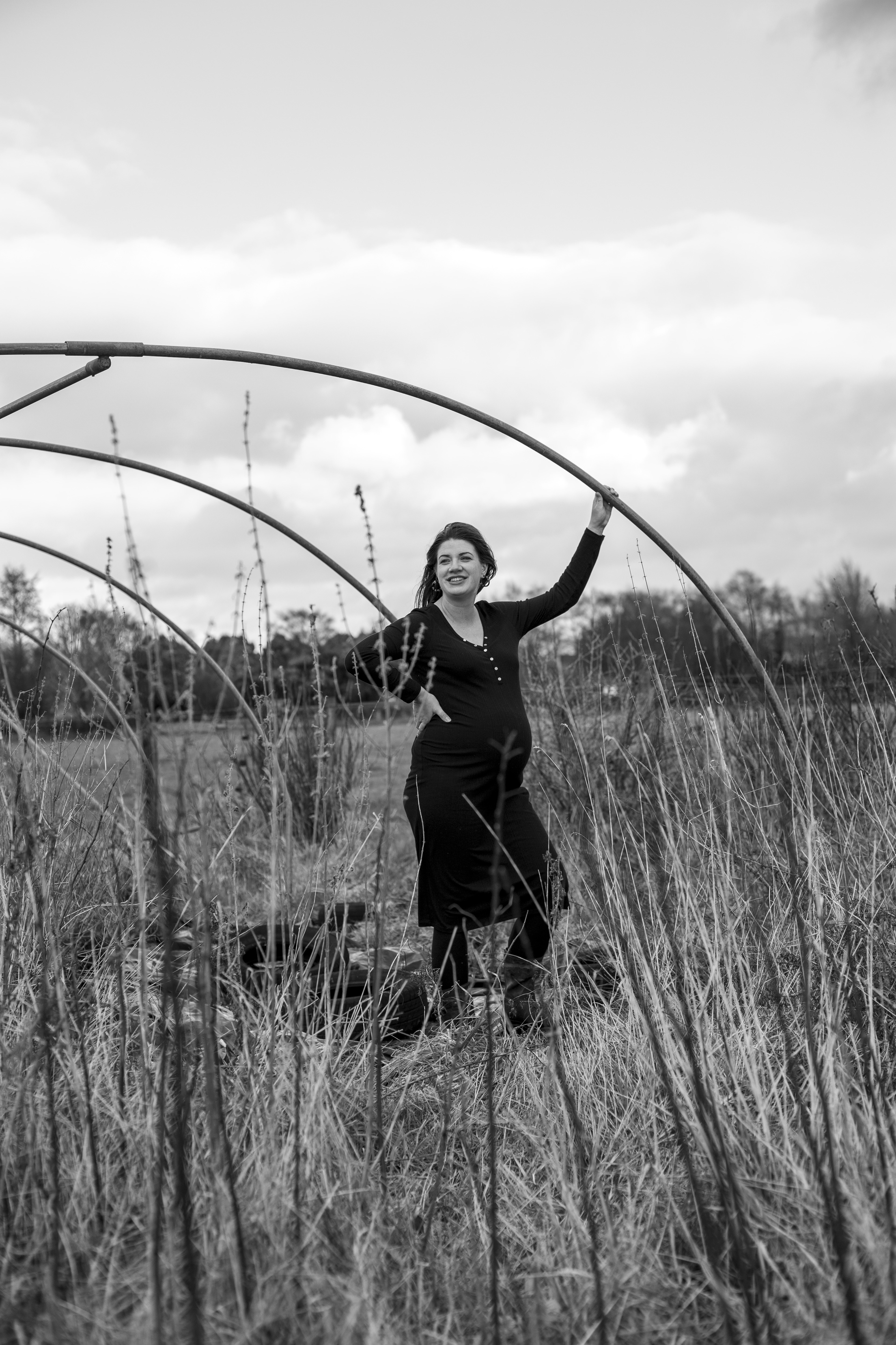 B&W portrait of pregnant woman holding polytunnel pole in grassy field