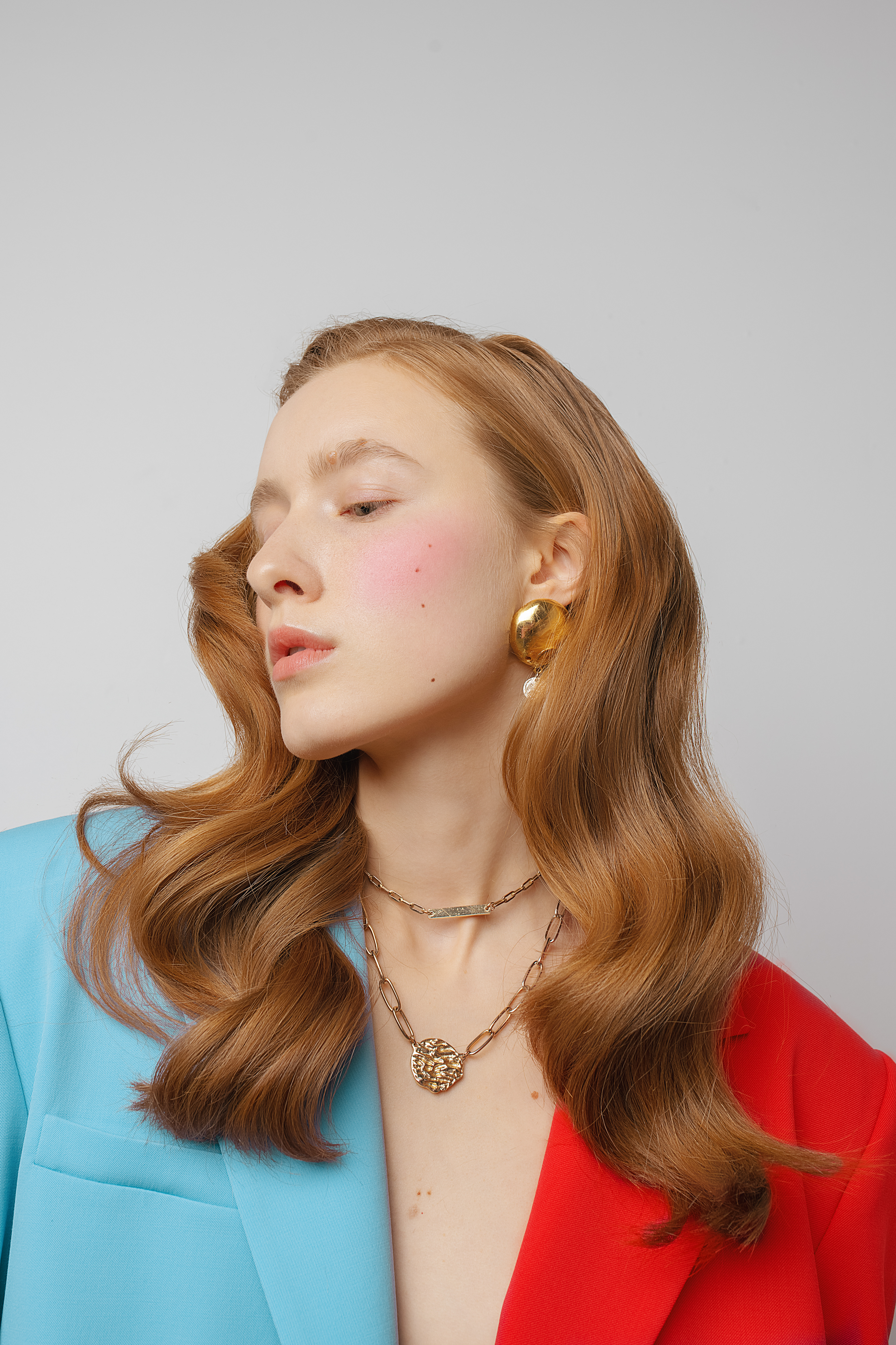 editorial shot of a woman in a red and cyan blazor, wearing long ginger head and gold jewelry.