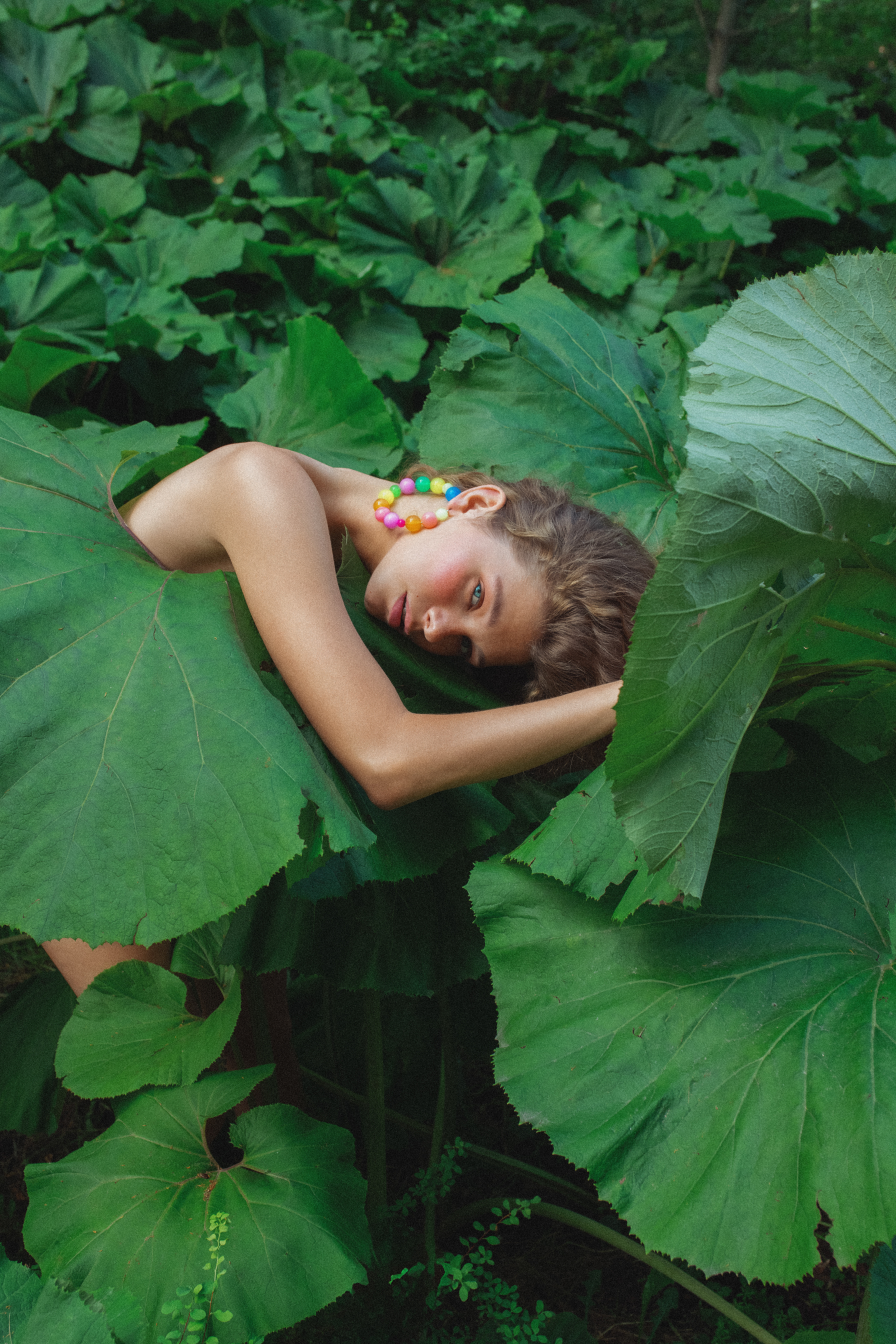 shot of a woman in a green forest, leaning on a big green leaf. Wearing a big circular rainbow earring.