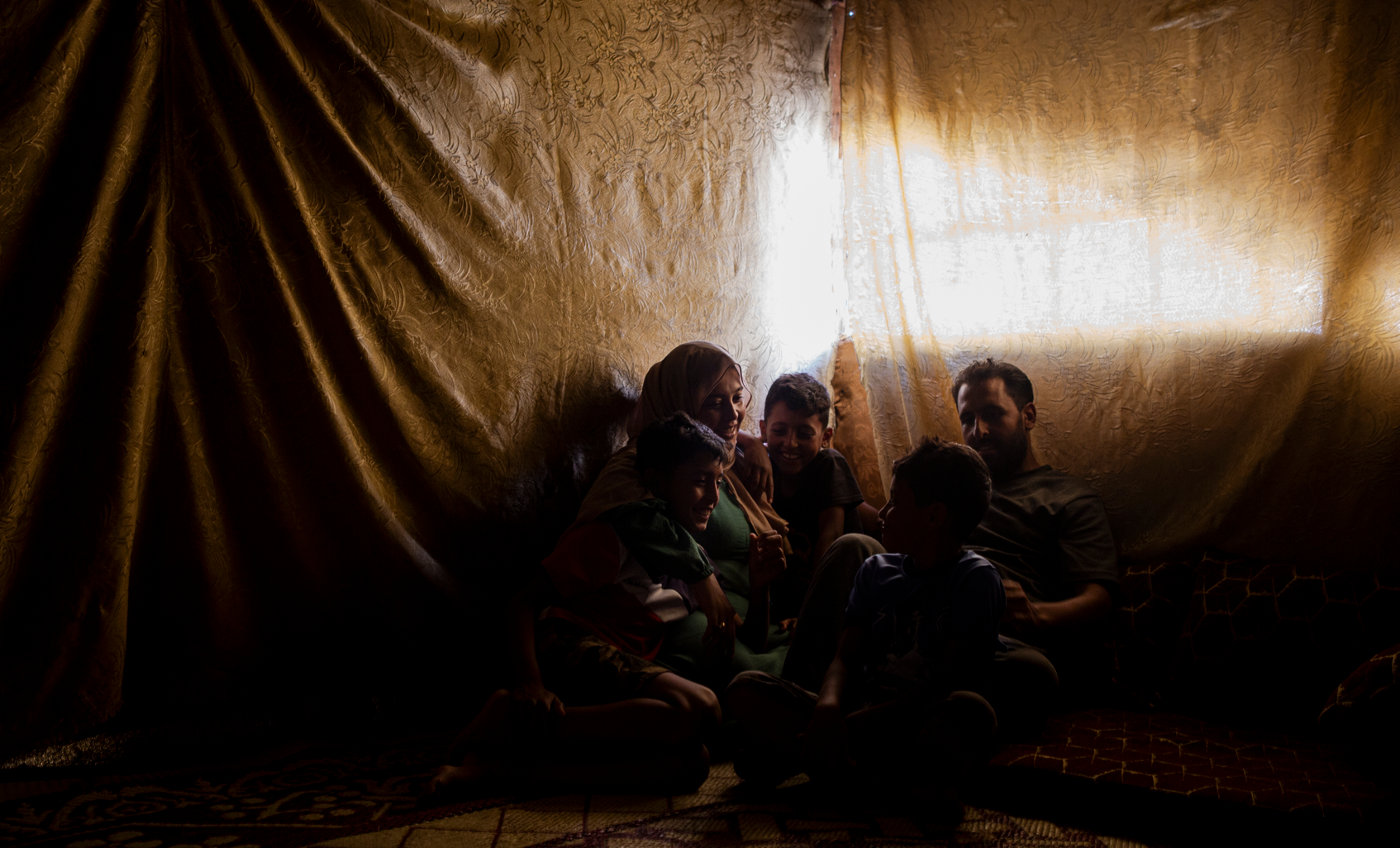 Bushra and her family sit for a portrait in their tented shelter. The walls are covered in gold fabric. Licing in this tented settlement in Lebanon for more than 12 years, Bushra has made this makeshift shelter a home for her and her family.
