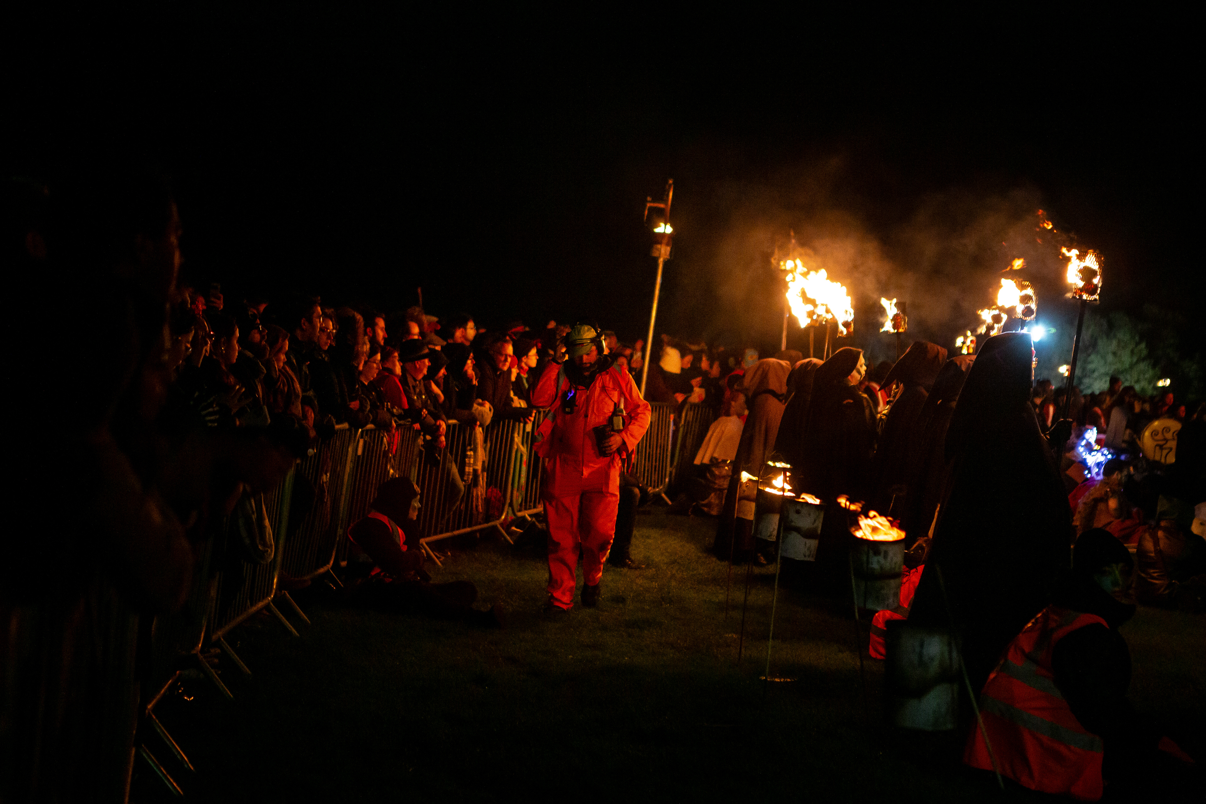 A safety person walk between the crowds behind a fence and performers holding lit torches at the festival.