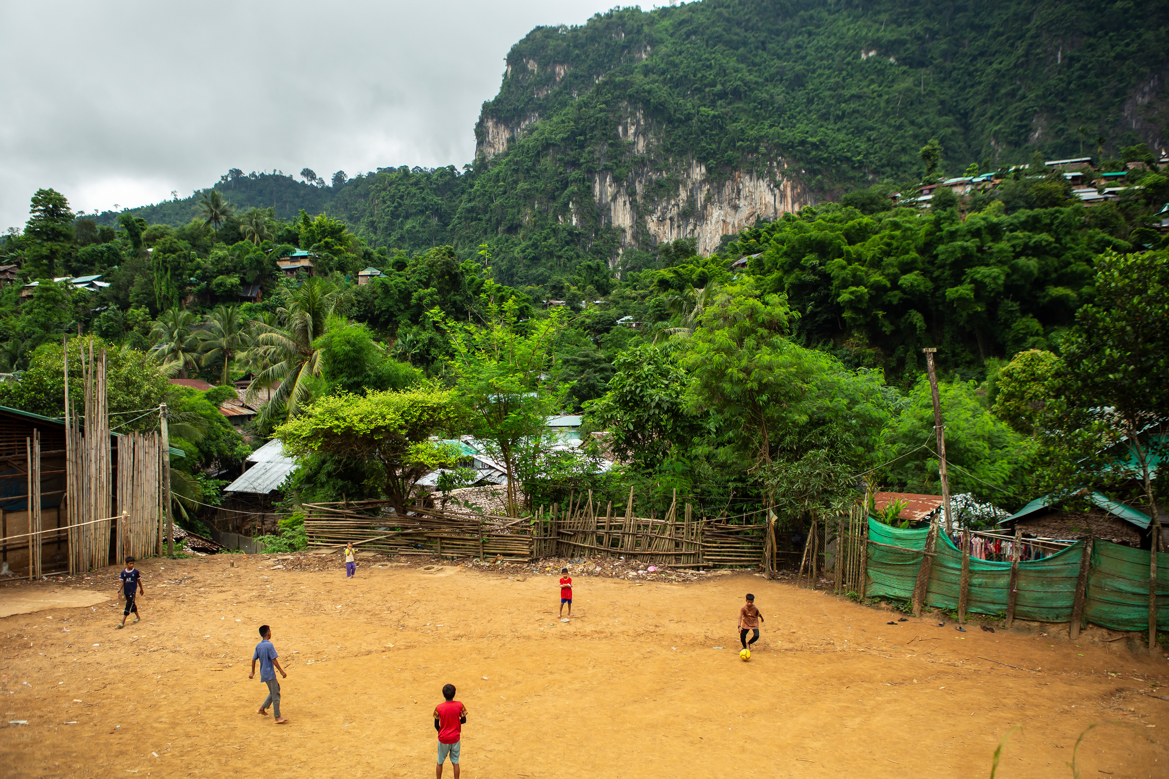 Some boys play football in the largest refugee camp in South East Asia, Mae-La refugee camp, which is estimated to house around 37,000 people and is located in Mae Sot near the border with Myanmar. Since the military coup which happened in Myanmar in 2021, a greater number of refugees have fled to Mae Sot as political violence has escalated throughout Myanmar. Refugees who live in this camp are guarded by the Thai military and confined to the camp, threatened with fines if they are found by the Thai military outside. July 2024