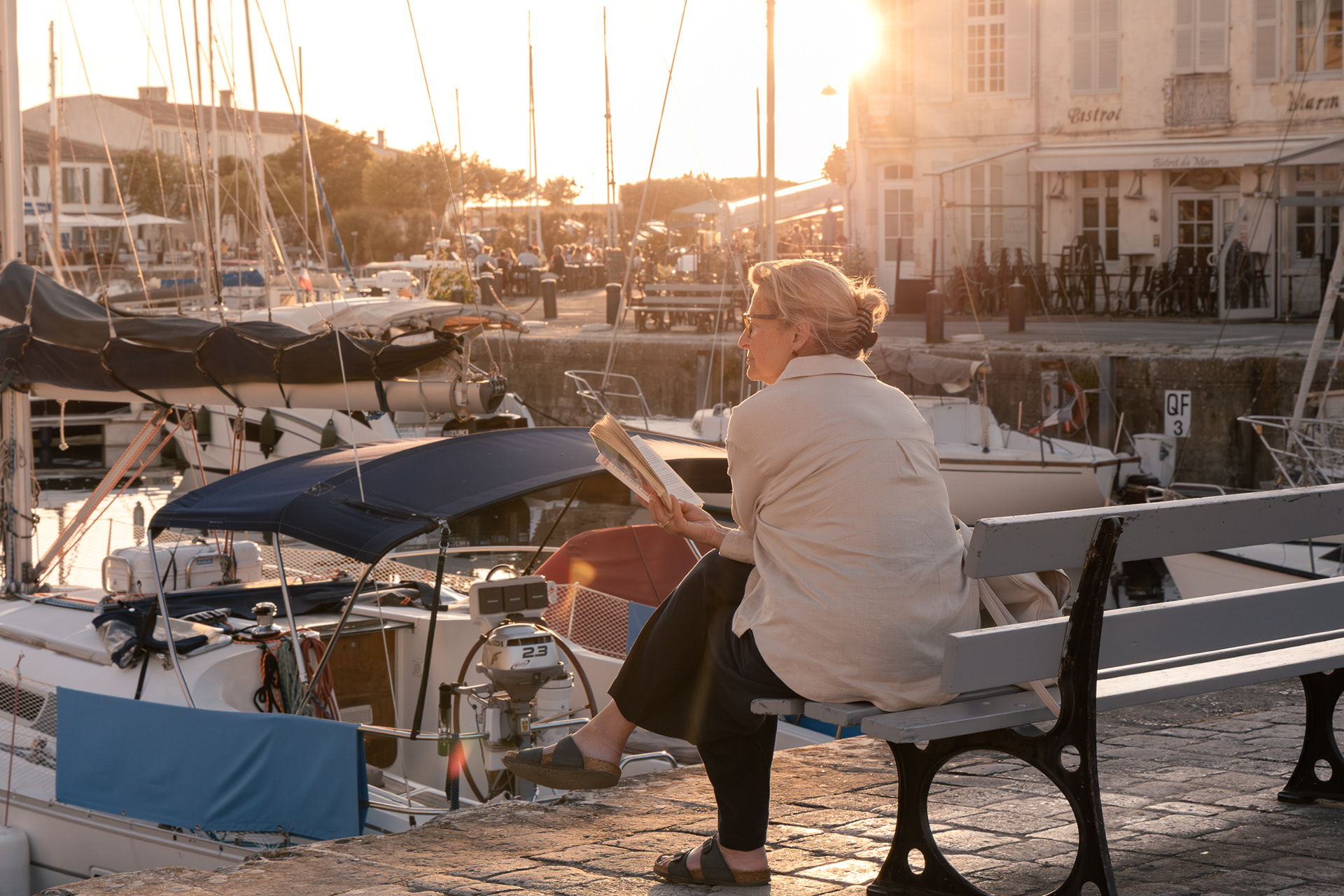 Street photography shot, framing a stranger lost in a book, contrasted against the vibrant evening energy around the harbour during golden hour.