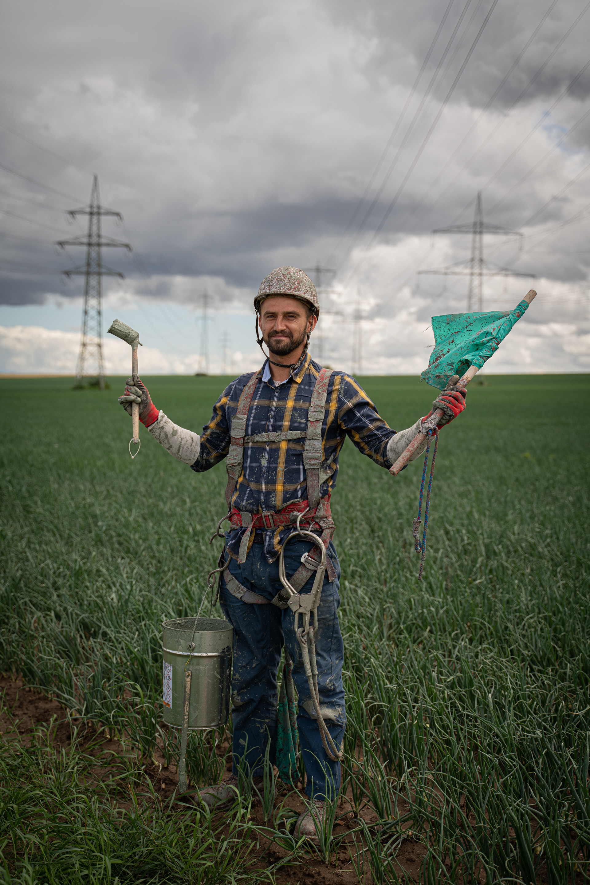 David, ein polnischer Arbeiter, der die Hochspannungsleitungen über einer Allee mit Rostschutzfarbe lackiert (Rheinland-Pfalz). Mit der grünen Fahne deutet er seinem Kol- legen, wie der Wind steht.