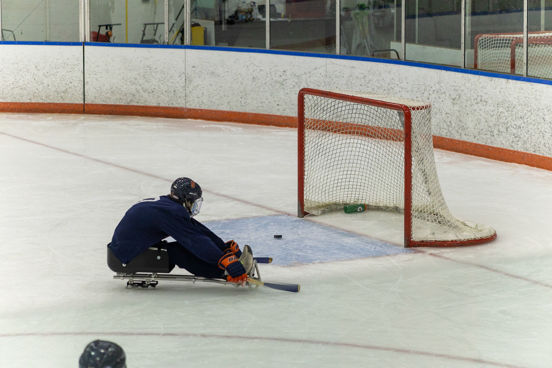 The CNY Flyers, a sled hockey team, played against the Syracuse University Men's Club Hockey team on 11/19/22 at Tennity Ice Skating Pavilion. The game ended with the Flyers scoring 8 to Syracuse 0 with 25 shots on goal by the Flyers compared to Syracuse's 2. While the game was still competitive, it was more for the fun of the sport with smiles shared by all. It was also a new experience for the Syracuse team which consisted of able-bodied individuals who may have never experienced sled hockey. The Syracuse goalie is absent from the goal as a hit from the Flyers scrapes just past the goal due to a small block by the Syracuse player in frame.