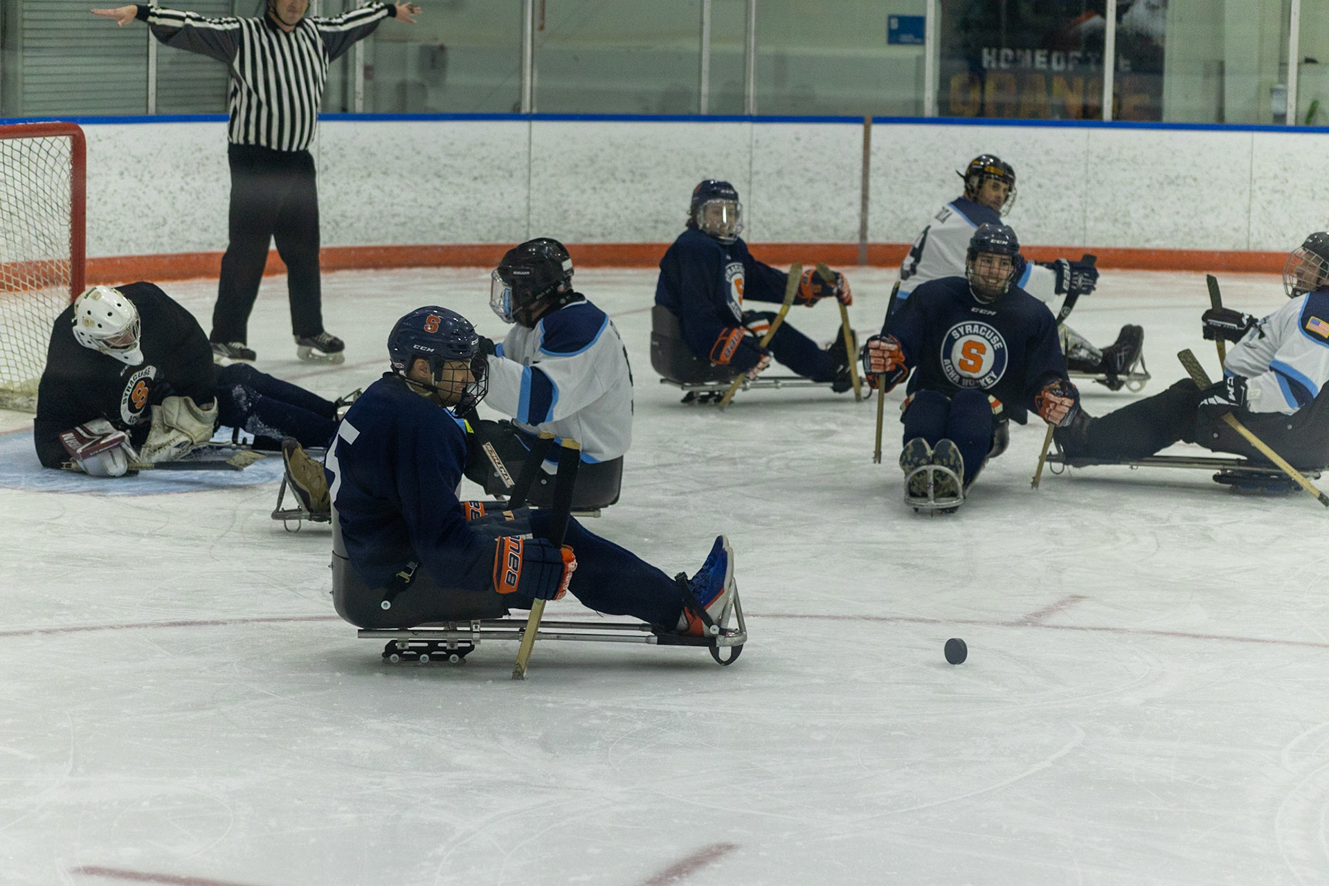 The CNY Flyers, a sled hockey team, played against the Syracuse University Men's Club Hockey team on 11/19/22 at Tennity Ice Skating Pavilion. The game ended with the Flyers scoring 8 to Syracuse 0 with 25 shots on goal by the Flyers compared to Syracuse's 2. While the game was still competitive, it was more for the fun of the sport with smiles shared by all. It was also a new experience for the Syracuse team which consisted of able-bodied individuals who may have never experienced sled hockey. The referee, Garth Werner, calls no goal as the puck spins across the rink.