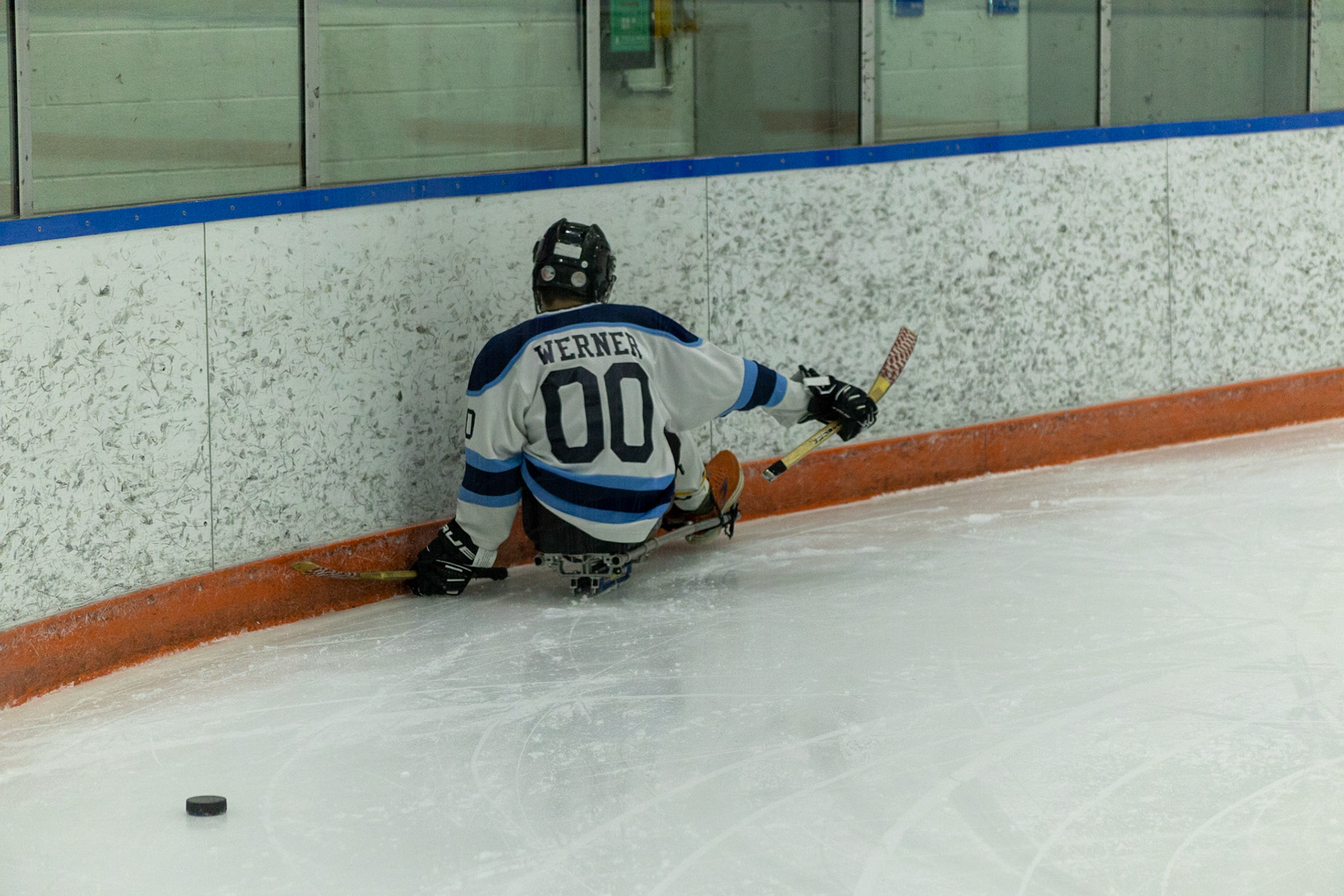 The CNY Flyers, a sled hockey team, played against the Syracuse University Men's Club Hockey team on 11/19/22 at Tennity Ice Skating Pavilion. The game ended with the Flyers scoring 8 to Syracuse 0 with 25 shots on goal by the Flyers compared to Syracuse's 2. While the game was still competitive, it was more for the fun of the sport with smiles shared by all. It was also a new experience for the Syracuse team which consisted of able-bodied individuals who may have never experienced sled hockey. #00, Ryan Werner, attempts to recover from hitting the wall as the puck slides behind.