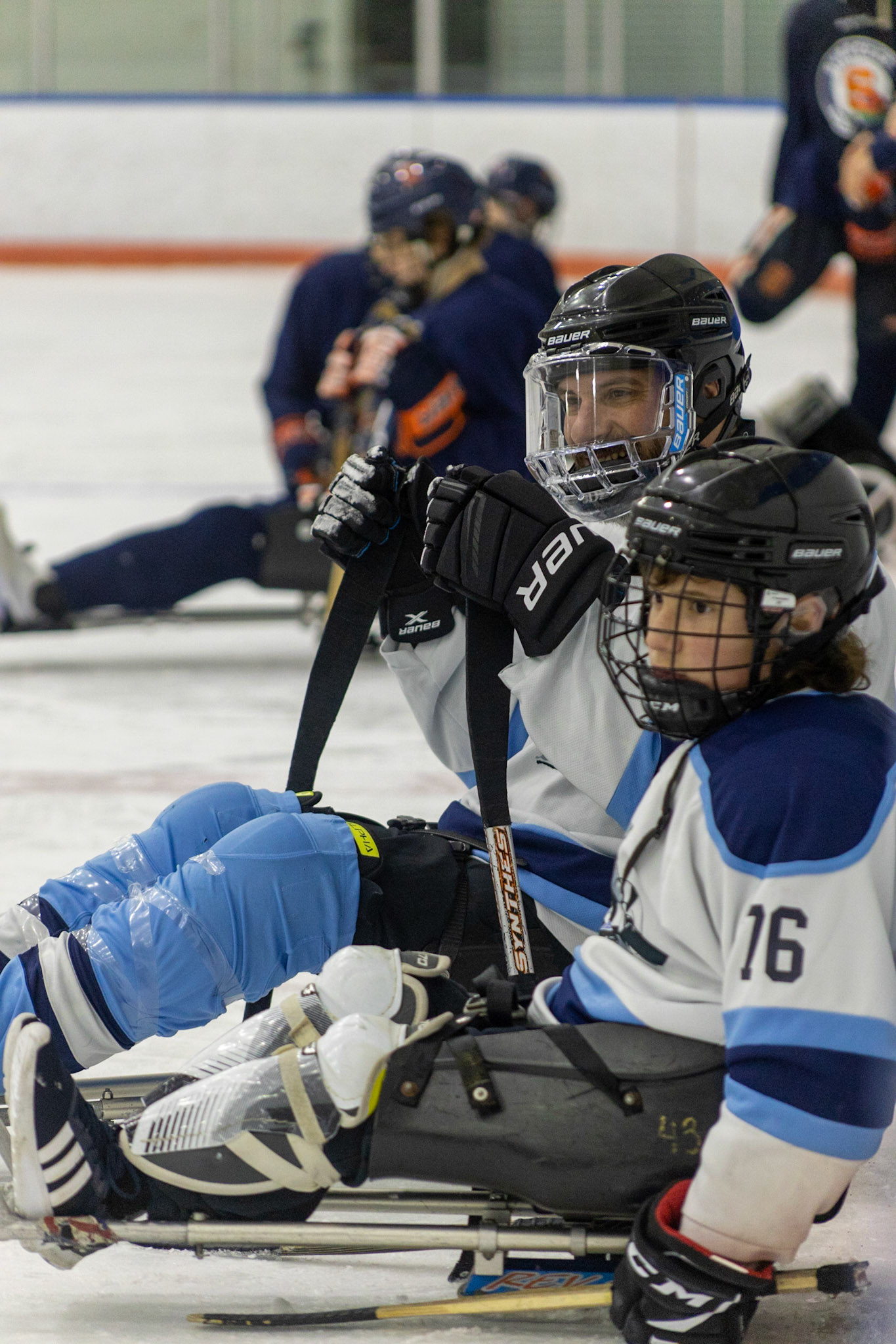 The CNY Flyers, a sled hockey team, played against the Syracuse University Men's Club Hockey team on 11/19/22 at Tennity Ice Skating Pavilion. The game ended with the Flyers scoring 8 to Syracuse 0 with 25 shots on goal by the Flyers compared to Syracuse's 2. While the game was still competitive, it was more for the fun of the sport with smiles shared by all. It was also a new experience for the Syracuse team which consisted of able-bodied individuals who may have never experienced sled hockey. #52 Seth Gitner and #76 Myles Favata rest just outside their teams bench. When it's time, they are tapped in by the team's coach who observes from within the box.
