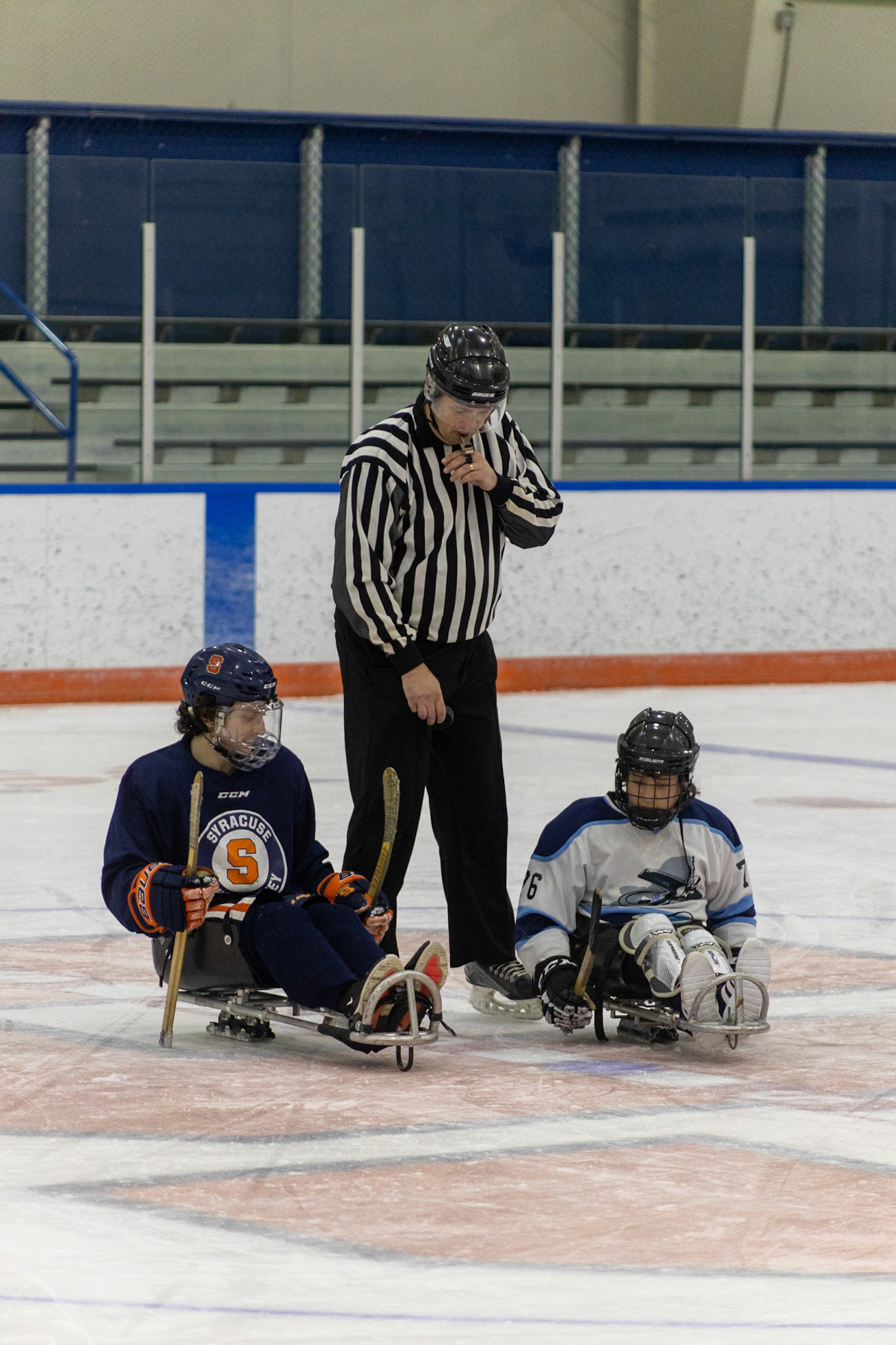 The CNY Flyers, a sled hockey team, played against the Syracuse University Men's Club Hockey team on 11/19/22 at Tennity Ice Skating Pavilion. The game ended with the Flyers scoring 8 to Syracuse 0 with 25 shots on goal by the Flyers compared to Syracuse's 2. While the game was still competitive, it was more for the fun of the sport with smiles shared by all. It was also a new experience for the Syracuse team which consisted of able-bodied individuals who may have never experienced sled hockey. In a faceoff, referee drops the puck to a Syracuse player and Myles Favata, #76 for the Flyers, who will then compete for possession of the puck.