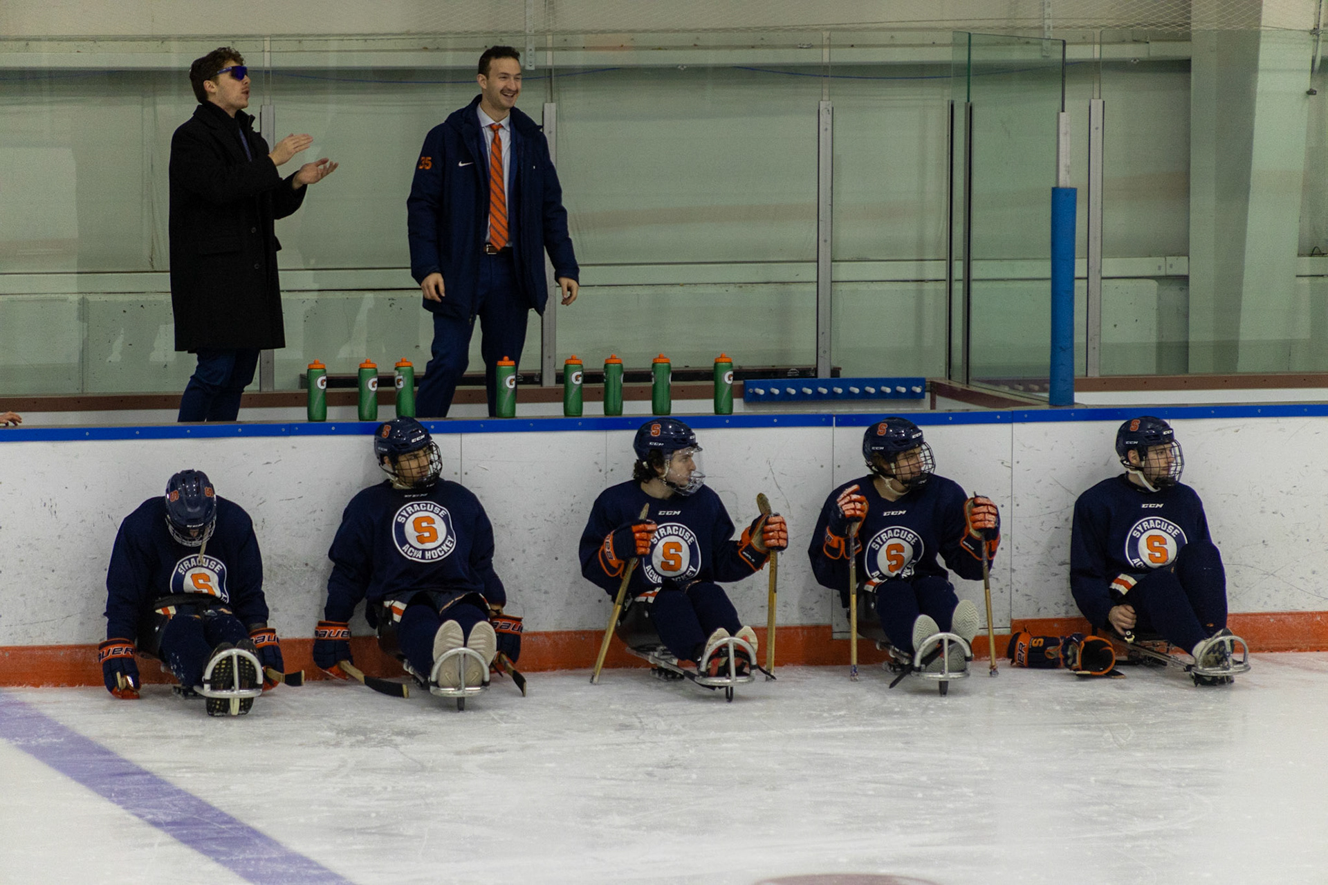 The CNY Flyers, a sled hockey team, played against the Syracuse University Men's Club Hockey team on 11/19/22 at Tennity Ice Skating Pavilion. The game ended with the Flyers scoring 8 to Syracuse 0 with 25 shots on goal by the Flyers compared to Syracuse's 2. While the game was still competitive, it was more for the fun of the sport with smiles shared by all. It was also a new experience for the Syracuse team which consisted of able-bodied individuals who may have never experienced sled hockey. Syracuse Hockey members sit in sleds by their box waiting to be tapped in by coaches. Above them, the coaches cheer on their team.