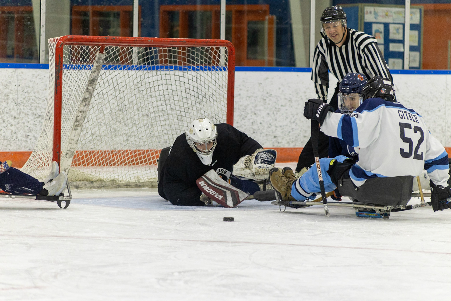The CNY Flyers, a sled hockey team, played against the Syracuse University Men's Club Hockey team on 11/19/22 at Tennity Ice Skating Pavilion. The game ended with the Flyers scoring 8 to Syracuse 0 with 25 shots on goal by the Flyers compared to Syracuse's 2. While the game was still competitive, it was more for the fun of the sport with smiles shared by all. It was also a new experience for the Syracuse team which consisted of able-bodied individuals who may have never experienced sled hockey. The Syracuse goalie dives to cover the goal as #52, Seth Gitner, attempts to reposition for an attempt on the goal.
