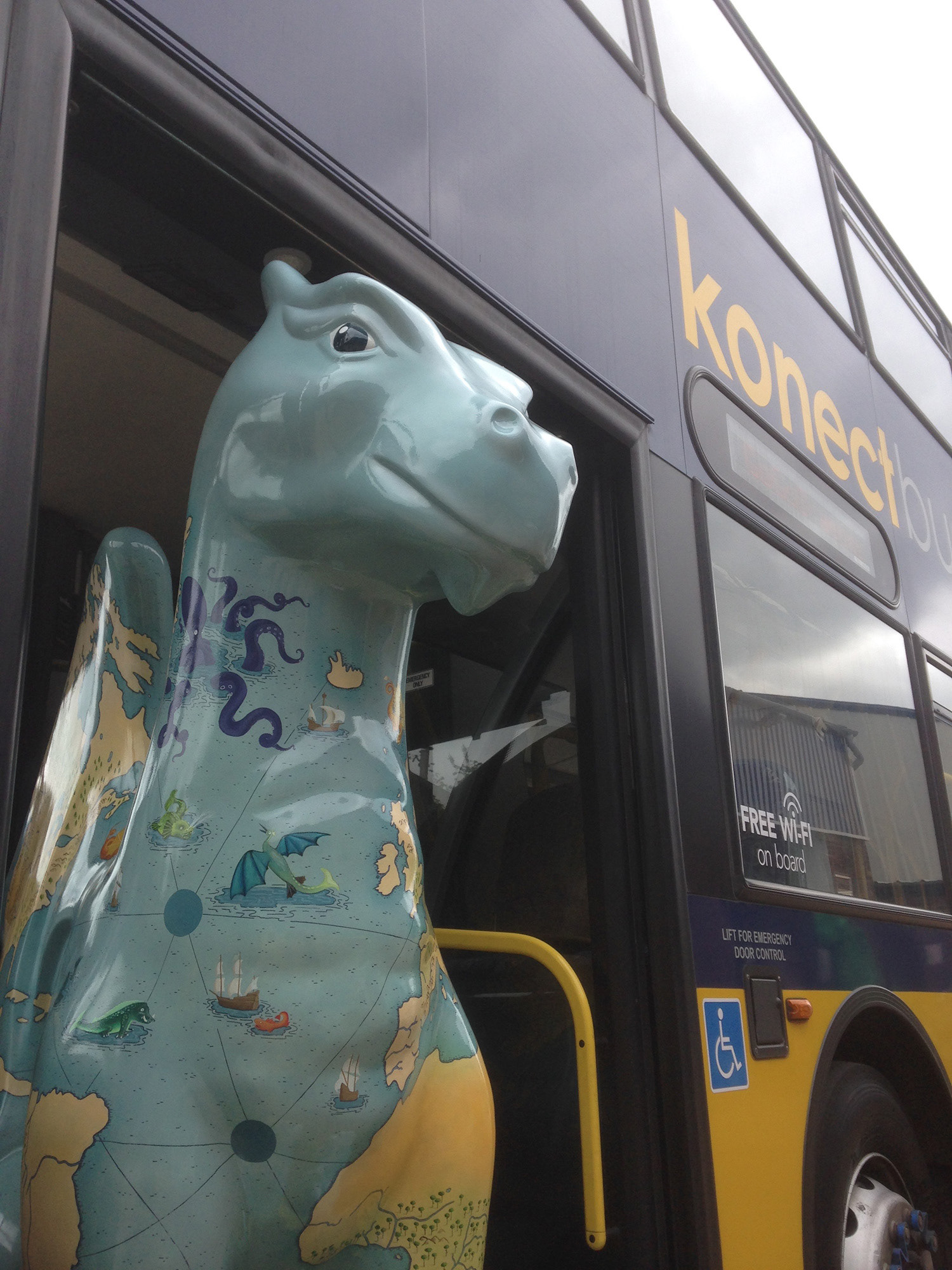 Dragon public art sculpture illustrated with a world map populated by sea monsters. Painted by Fiona Gowen. He is standing in a bus doorway.