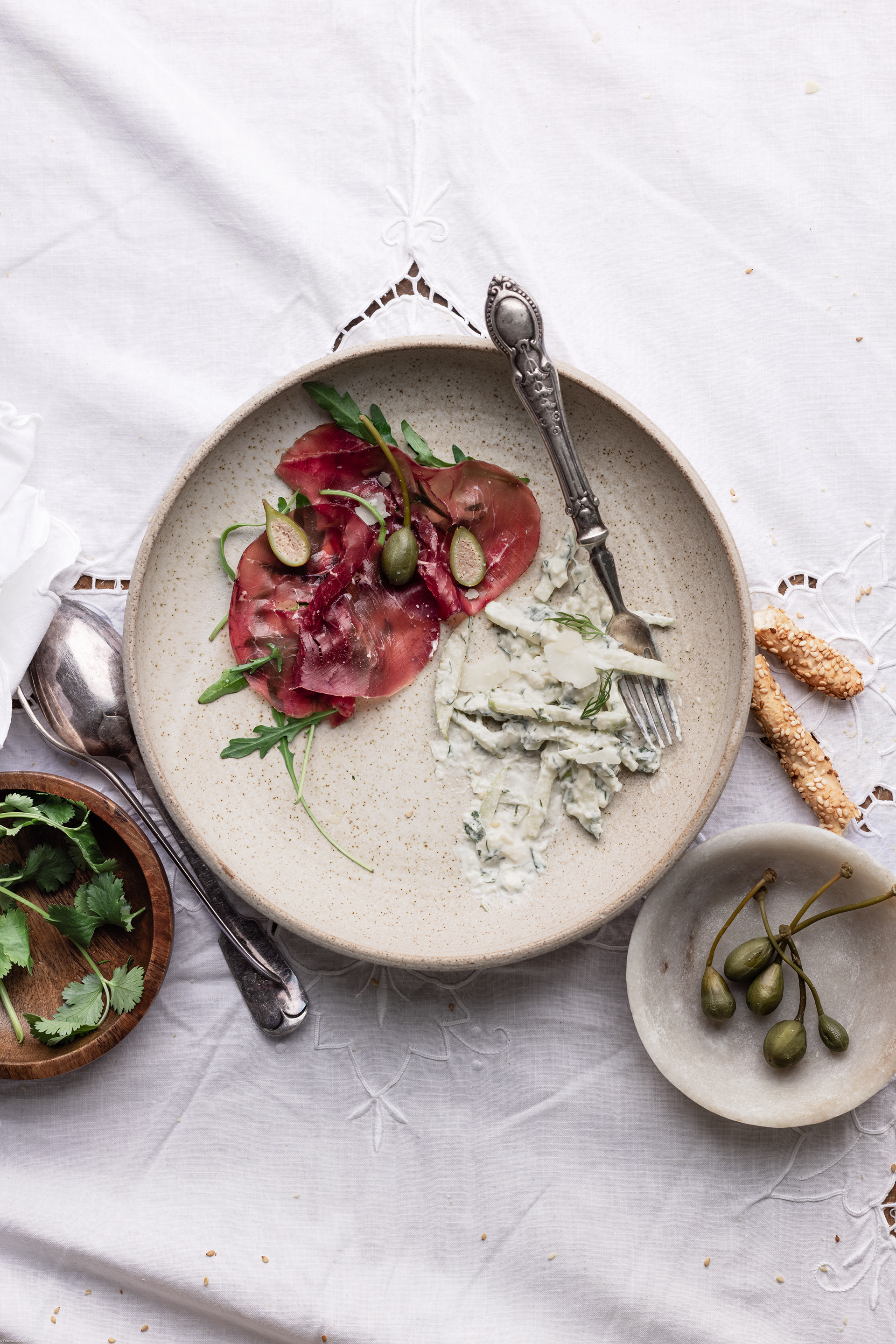 A food photography (fotografia di cibo) and food styling image of bresaola carpaccio with arugula and capers on a plate with some small bowls around made by Federica Melania Orsini - a food photographer (fotografo di cibo) in Bologna.