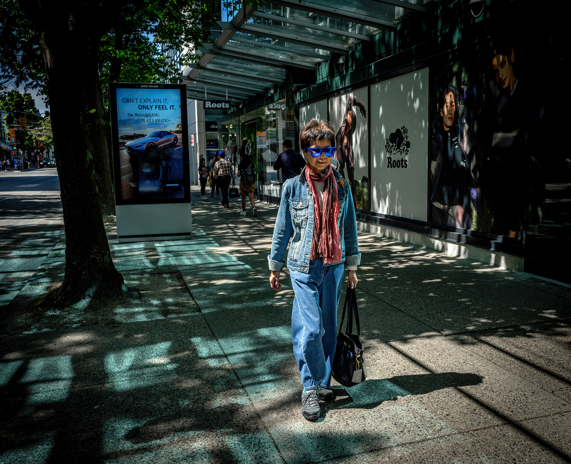 Woman wit blue glasses walking on a sidewalk, blue ONLY FEEL IT car advertisement behind her.