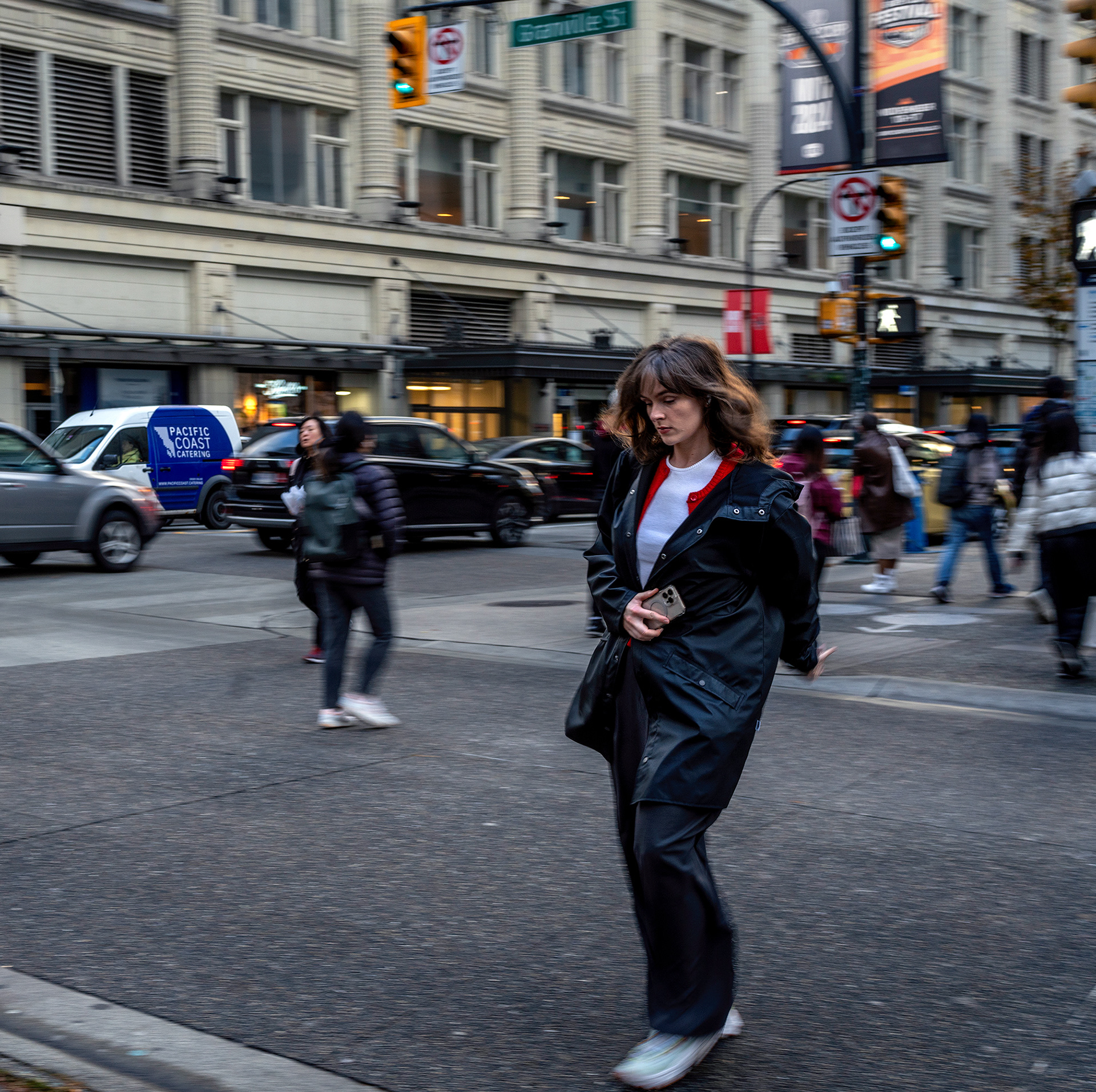 Bundled up woman ccrossing a sidewalk across the street from The Bay, in downtown Vancouver, she has her cellphone in hand.