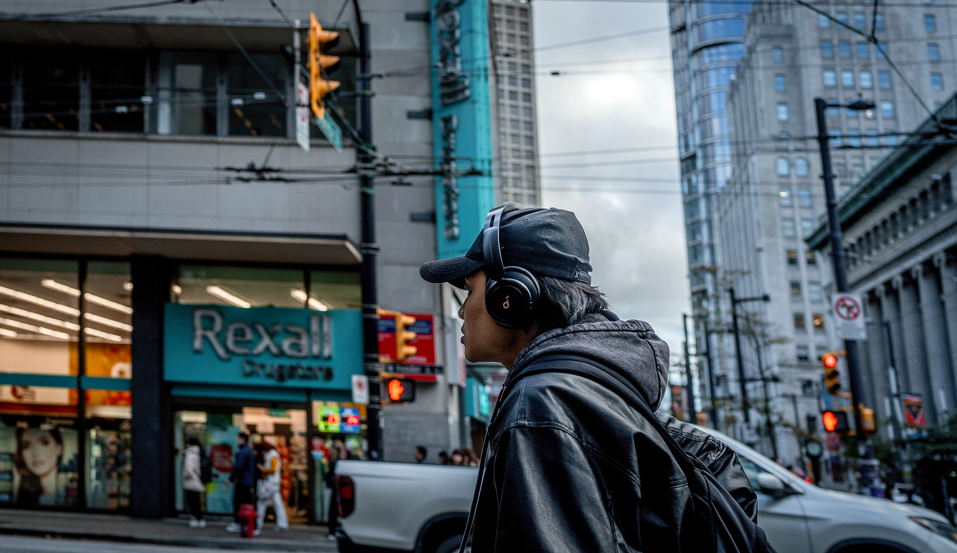 Man with black cap and headphones, across the street from a Rexaall pharmacy, downtown city scene.