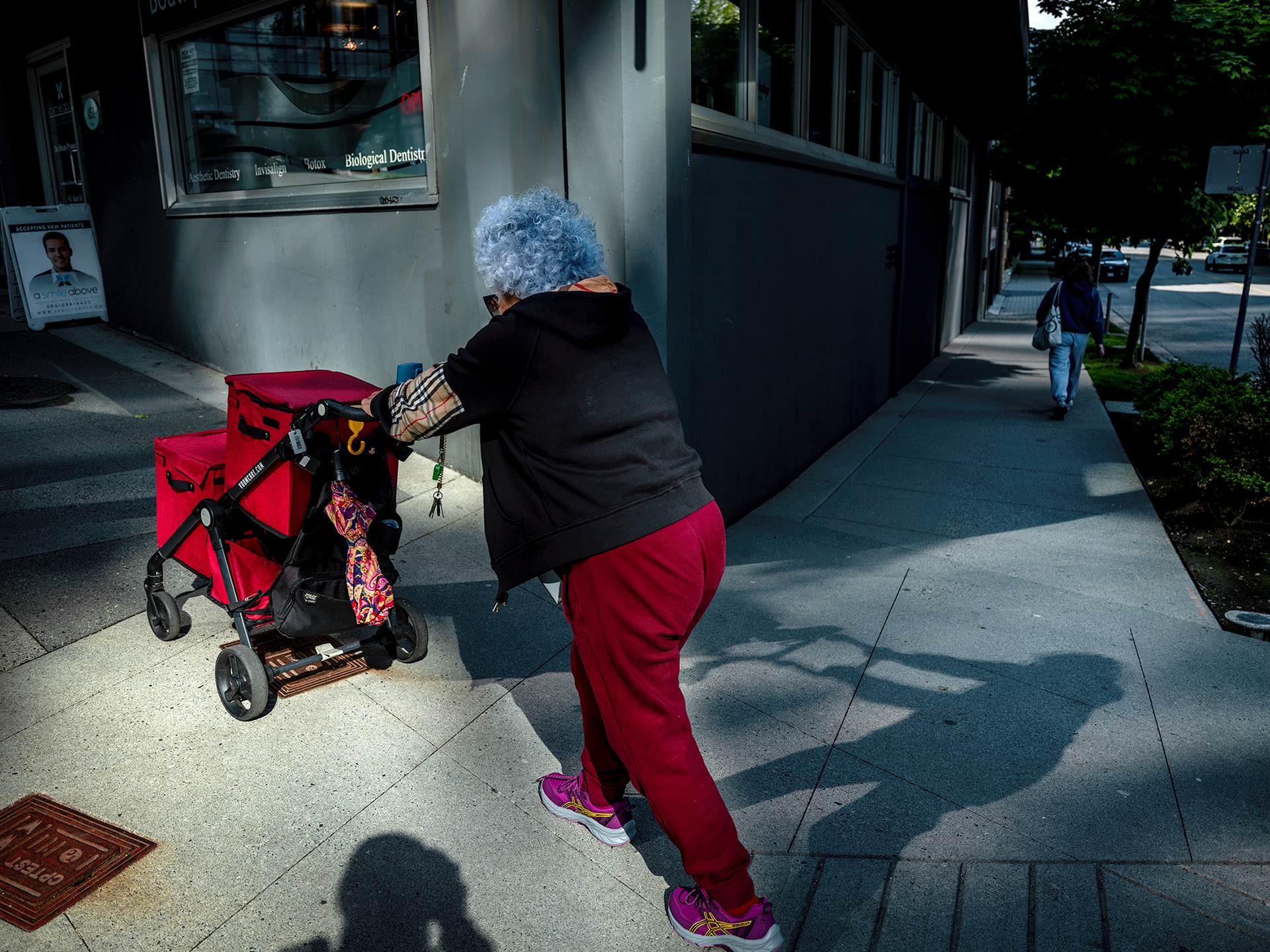 Woman with blue hair pushing heer red shopping cart up a steep sidewalk, dramatic lighting.