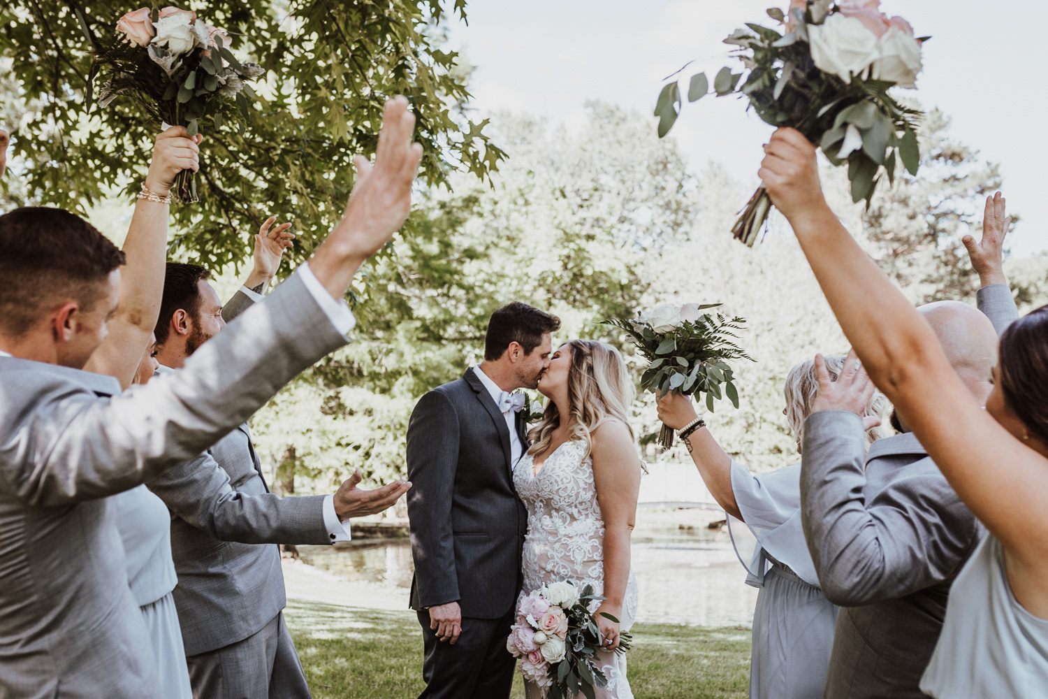 stylish bride and groom posing in front of small pond while wedding party stands on either side cheering for the couple in conway ar.